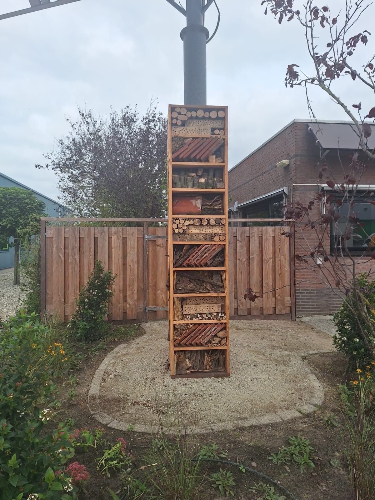 Hoog houten insectenhotel op maat, gevuld met stammetjes, dakpannen en takken, op een rond grindveld in de tuin