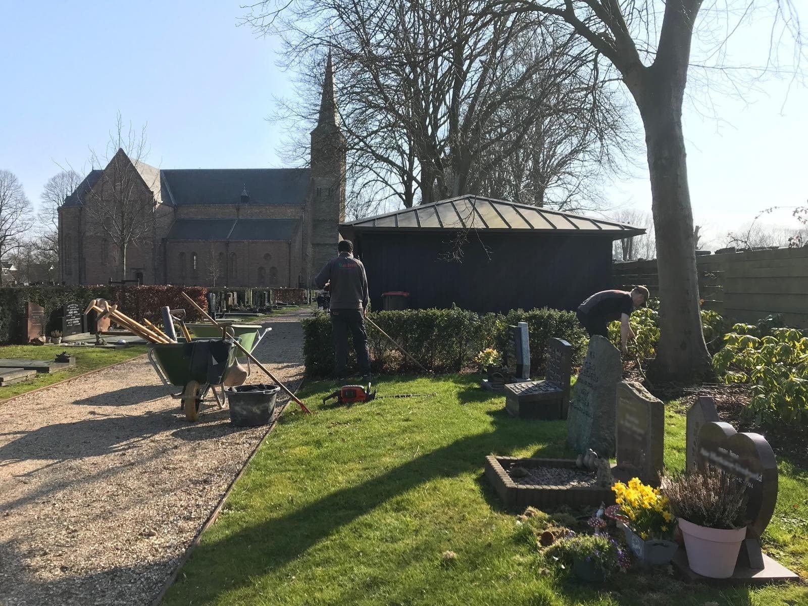 Twee medewerkers van Van Beets onderhouden de heesters en het gras tussen de graven op een dorpsbegraafplaats, met kruiwagens en gereedschap op het grindpad en de kerk op de achtergrond