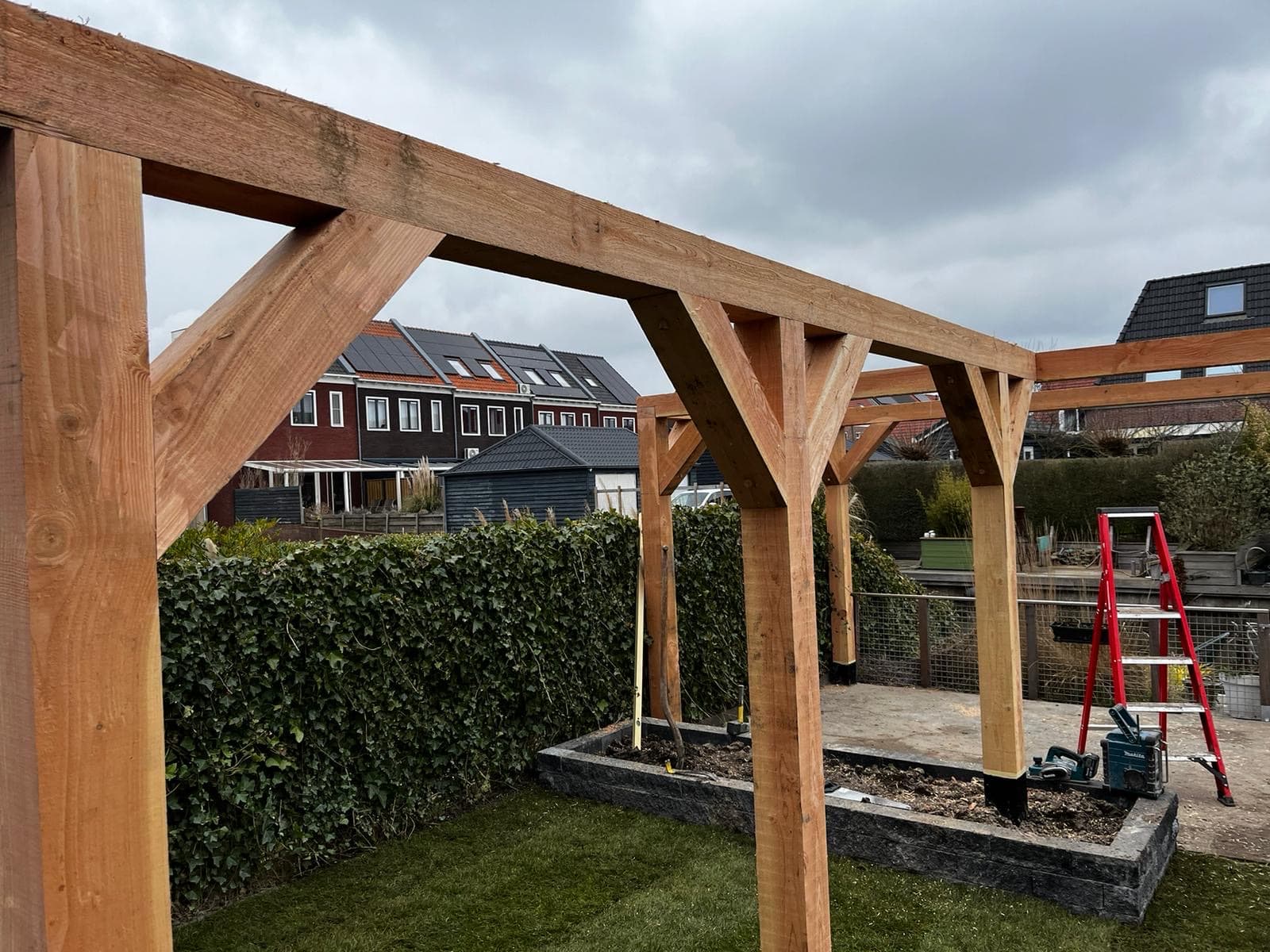Houten pergola van eikenhout met schoren boven een tuin met klimopheg, tijdens de bouw