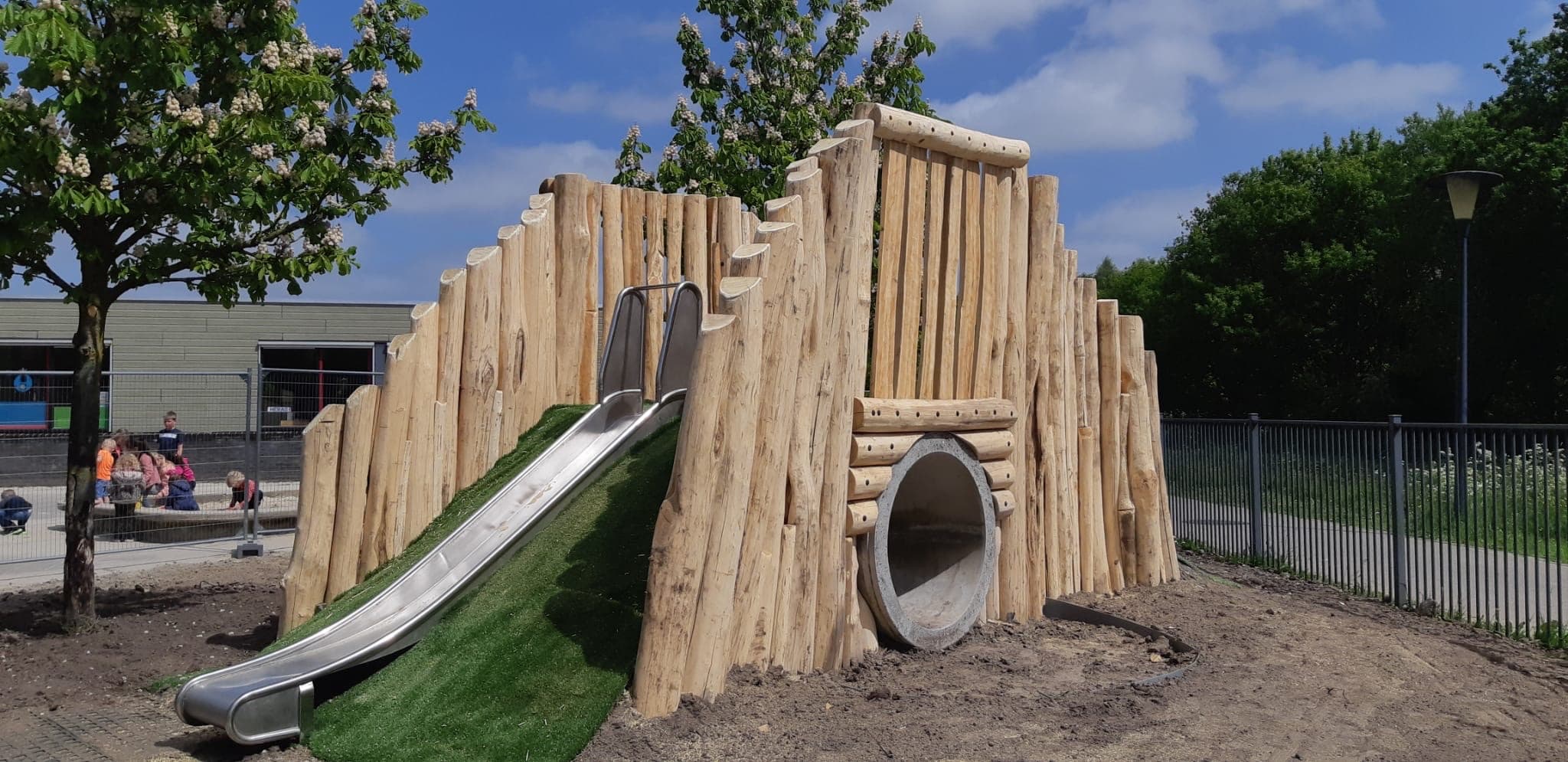 Houten speelheuvel van geschilde stammen met een roestvrijstalen glijbaan en een tunnel op een schoolplein