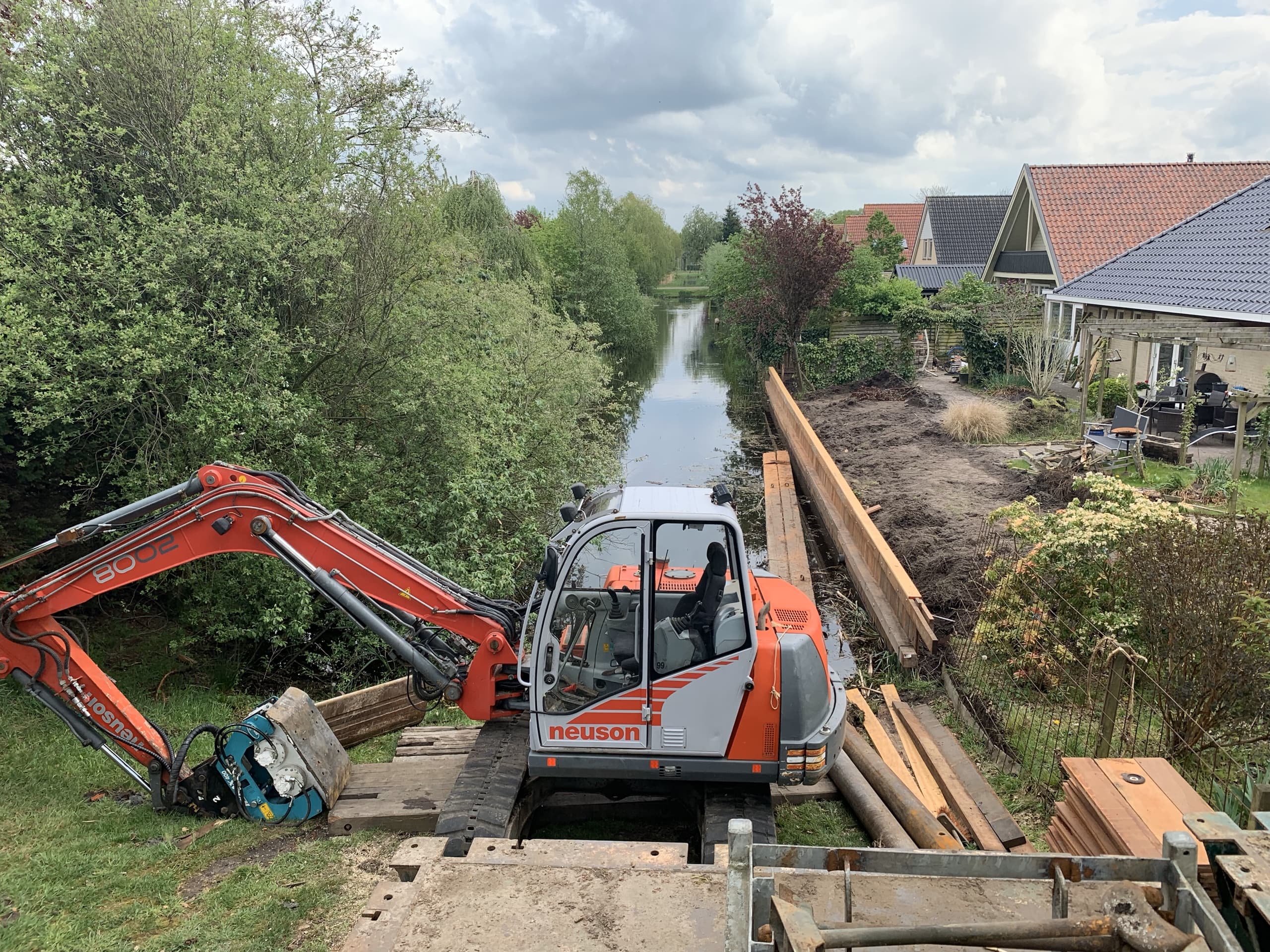 Rupskraan plaatst vanaf een tijdelijke werkbrug een nieuwe houten walbeschoeiing langs een tuin in Heerenveen