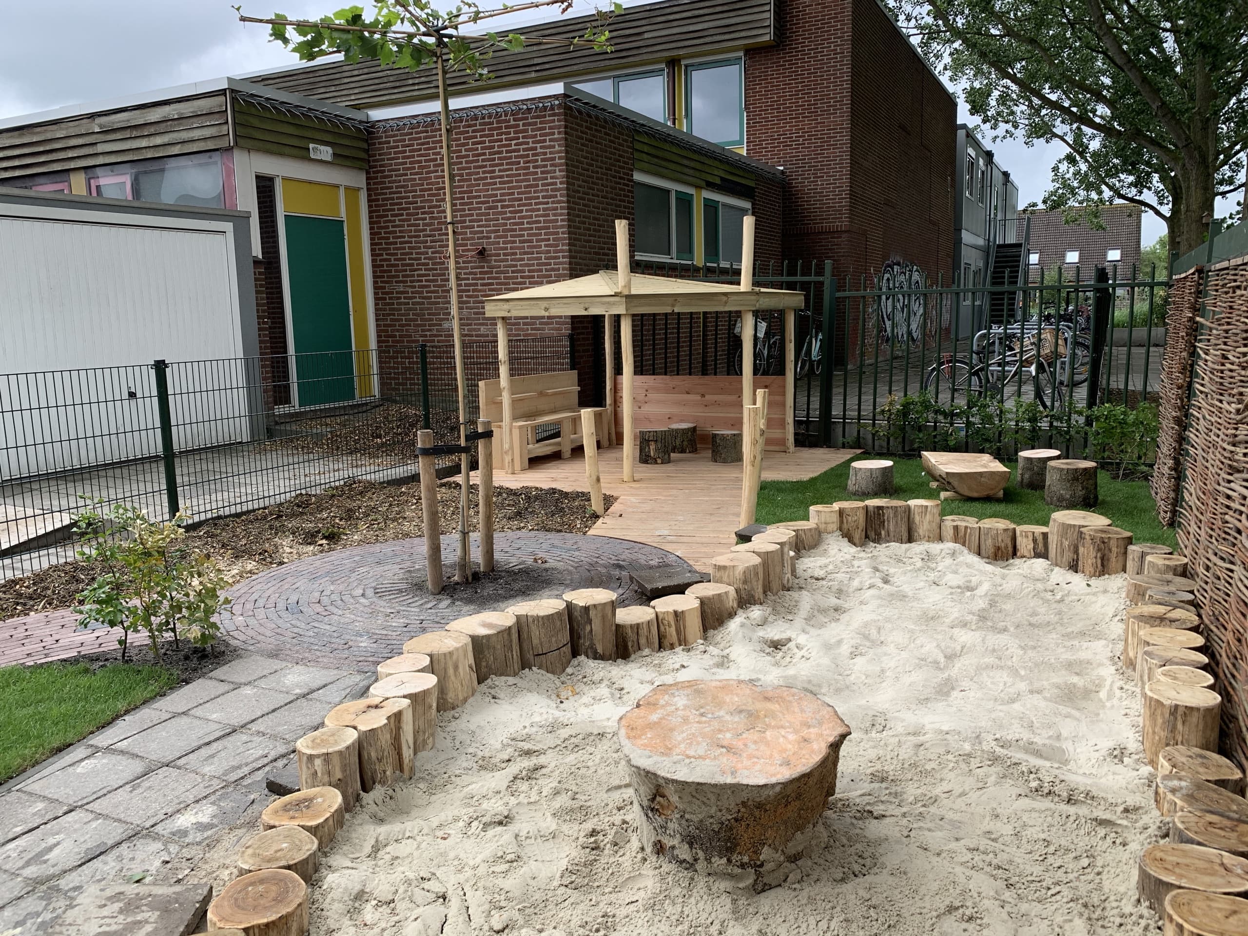 De speelplek met zandkuil omzoomd door boomstammen, een houten prieel met zitbank en een vlechtwerkscherm van wilgentenen
