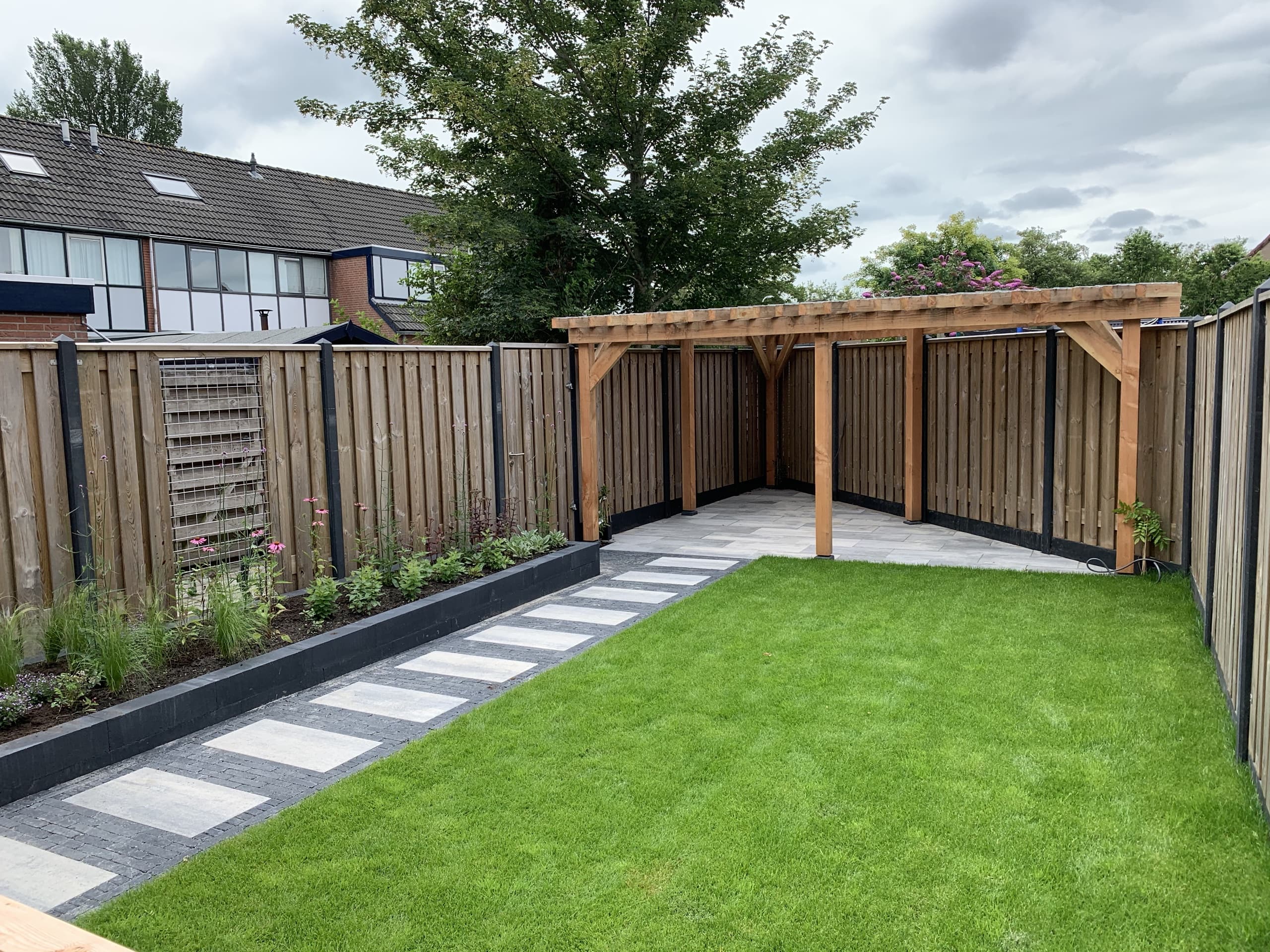 De volledige achtertuin met stapstenenpad, border, gazon en de houten pergola als zithoek achterin