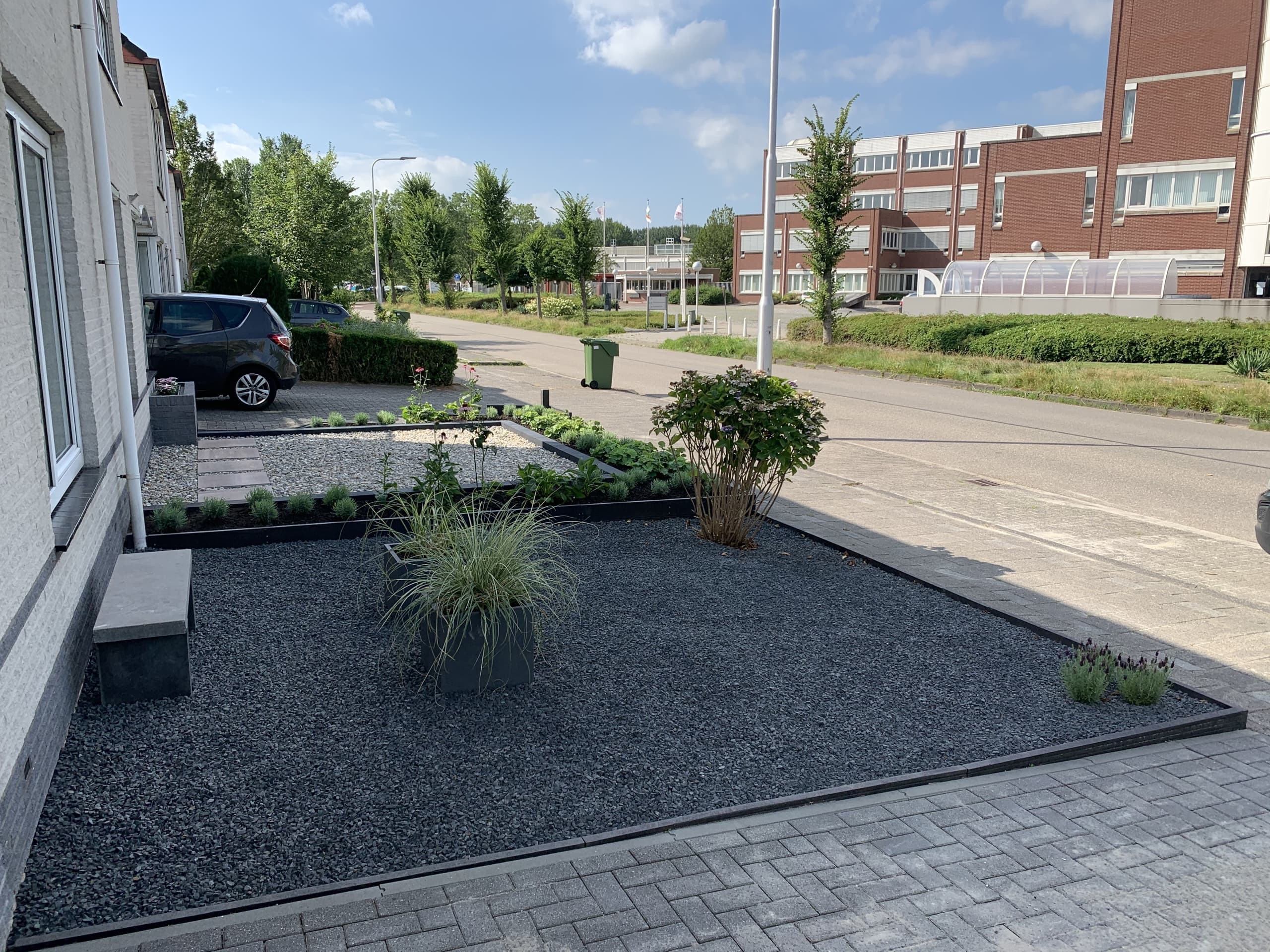 Het donkere grindvlak met plantenbakken van siergrassen en een hortensia, met de straat op de achtergrond