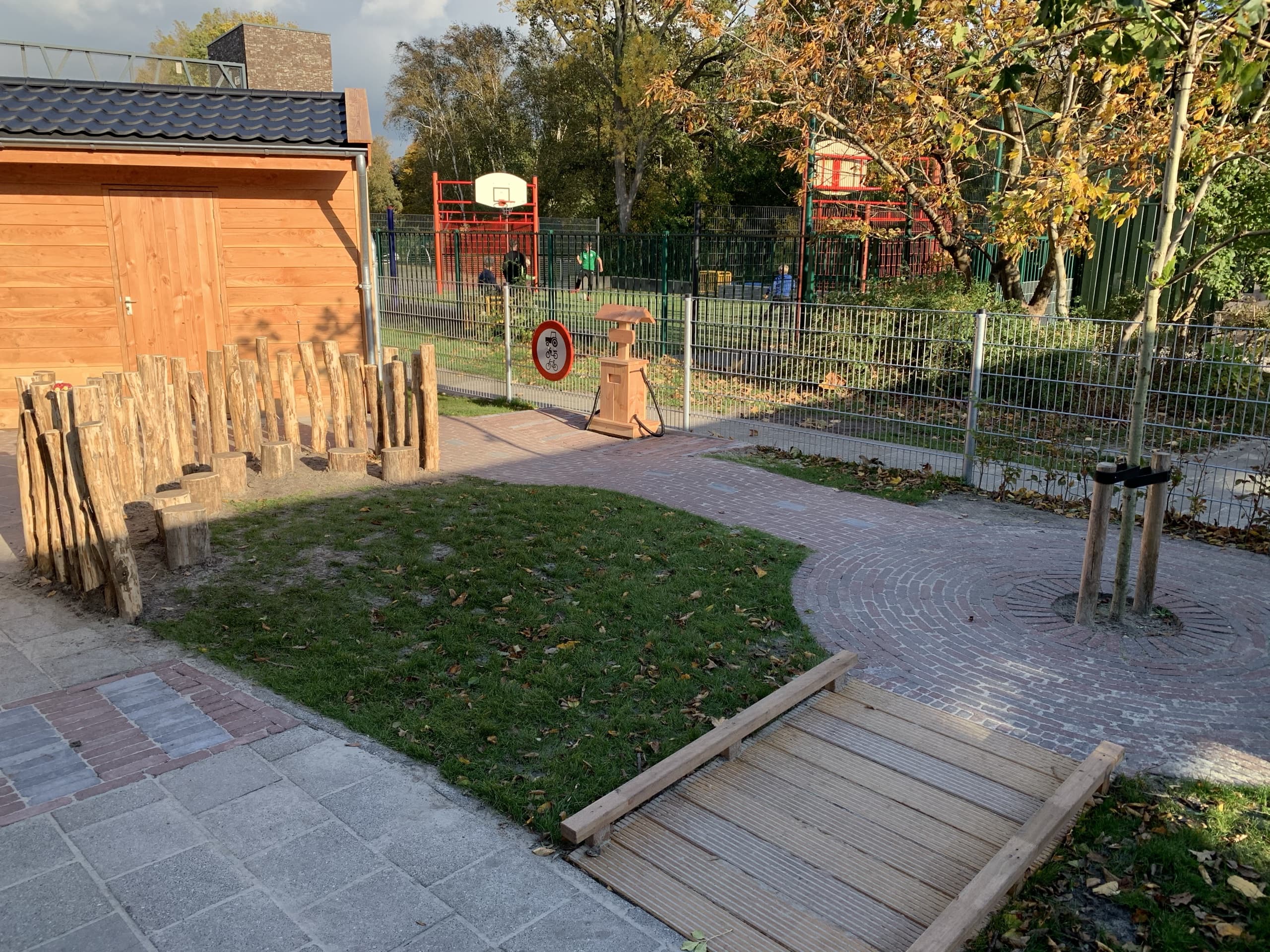 Een waterpomp op een houten sokkel naast een rij boomstammen als klimobstakel, met op de achtergrond het verharde plein met basketbalveld