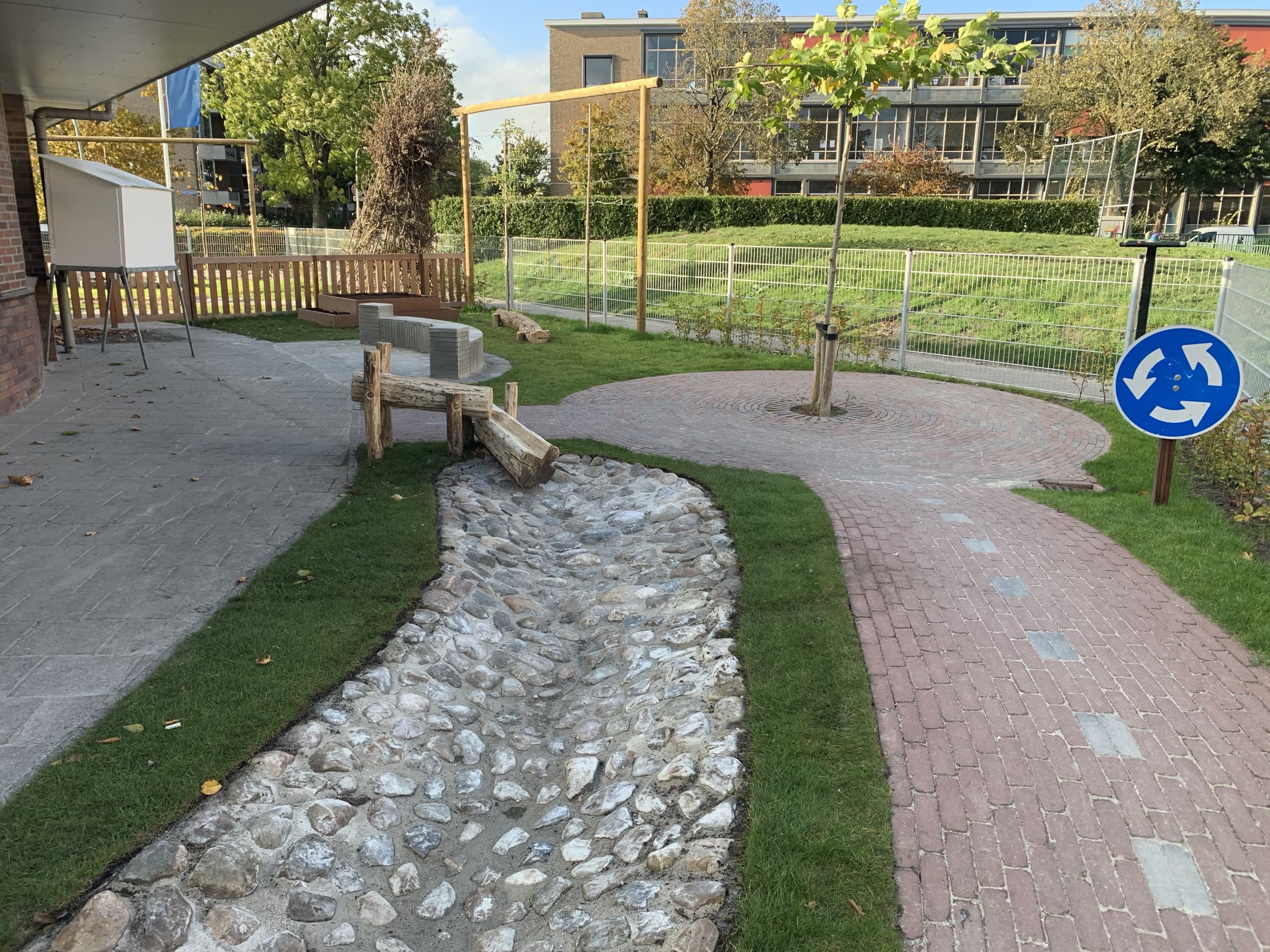Overzicht van de verkeerstuin: een kiezelbeek met houten duiker, een cirkelvormig bestratingspatroon met jonge boom en een rotondebord