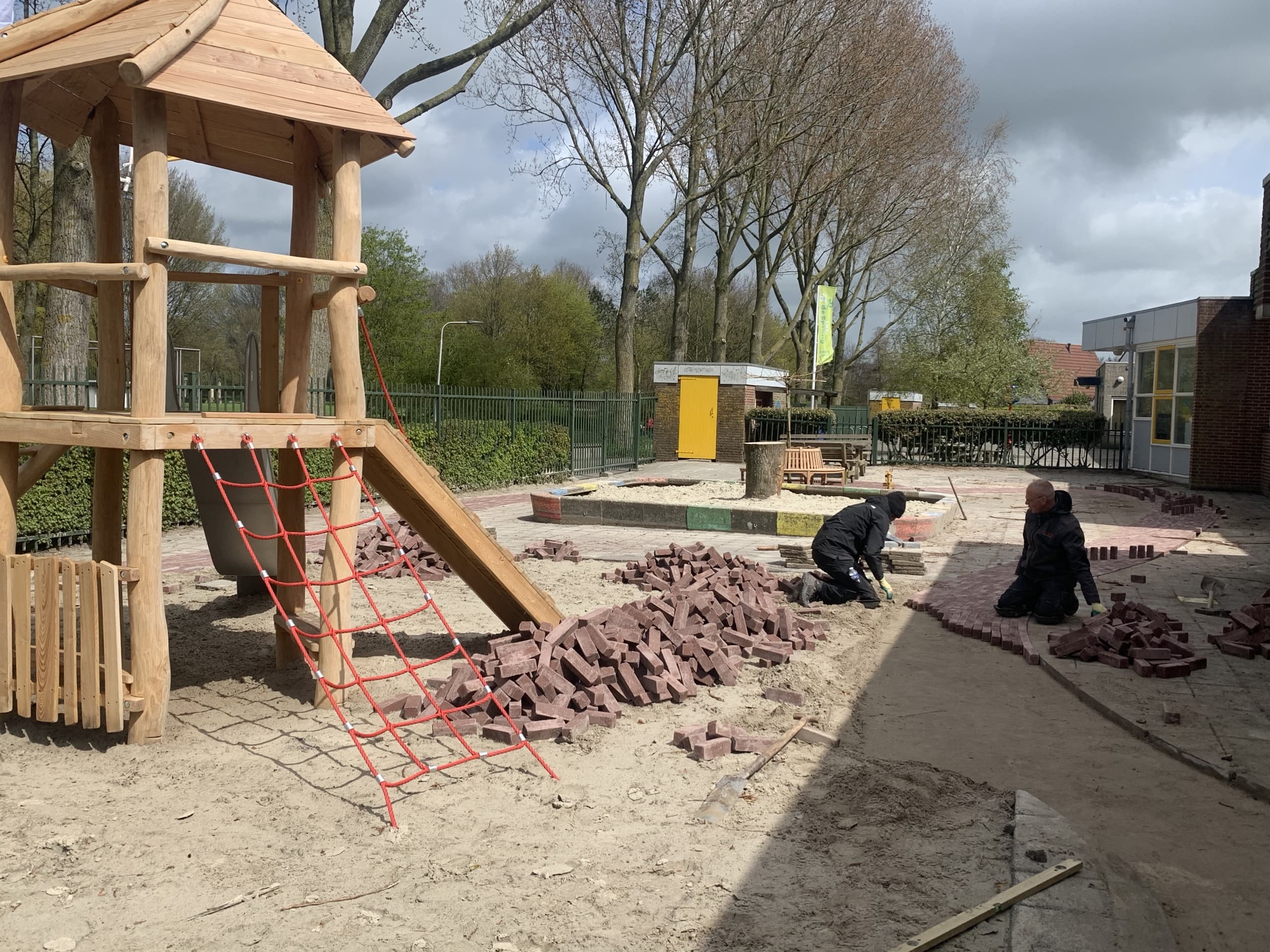 Twee stratenmakers van Van Beets leggen op hun knieën klinkers op een schoolplein, naast een houten speeltoestel