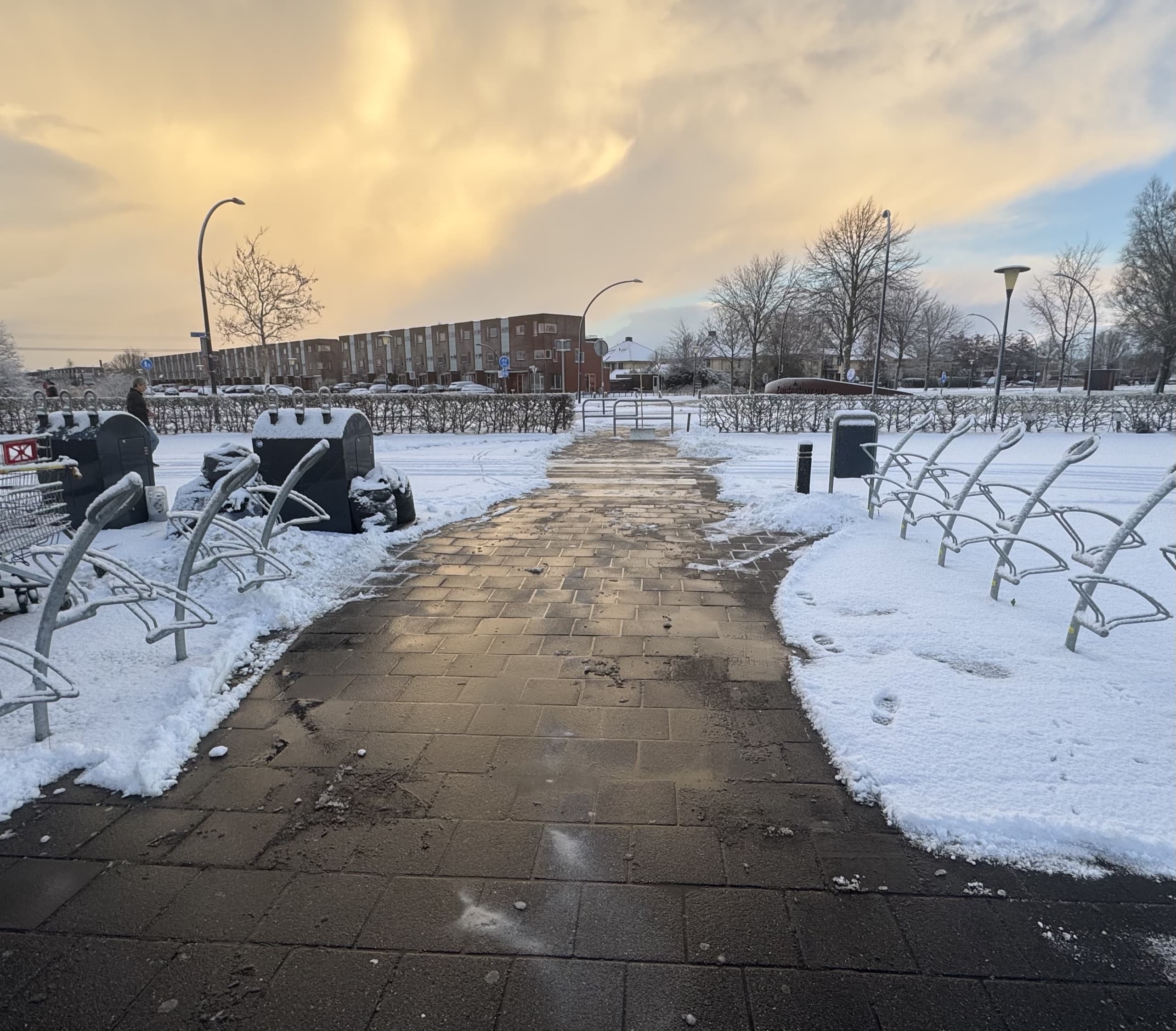 Sneeuwvrij geschoven voetpad tussen fietsenrekken bij een wooncomplex, met opzij geschoven sneeuw en een flatgebouw op de achtergrond bij zonsondergang
