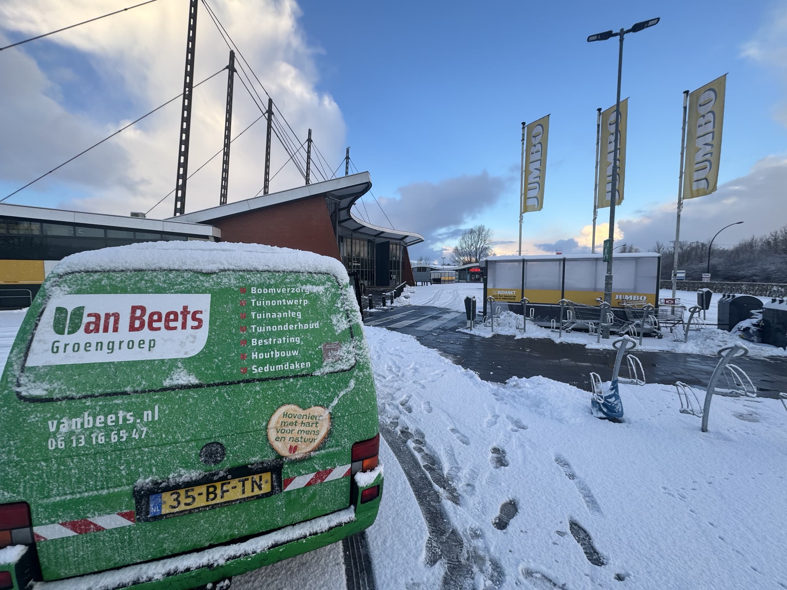 Besneeuwde bedrijfsbus van Van Beets Groengroep geparkeerd bij de ingang van een winkelcentrum met Jumbo-vlaggen, met sneeuwvrije paden en fietsenrekken op de achtergrond