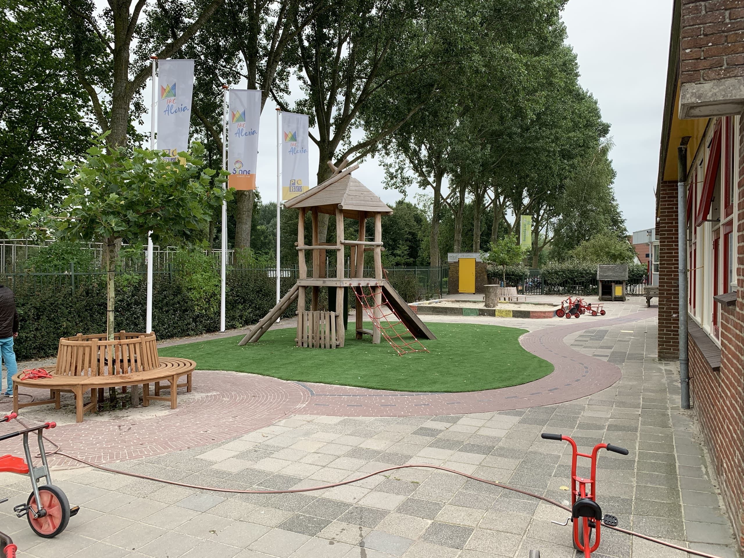 De klimtoren met glijbaan op het kunstgraseiland, met een gebogen houten bank om de boom en de vlaggen van IKC Alexia erachter