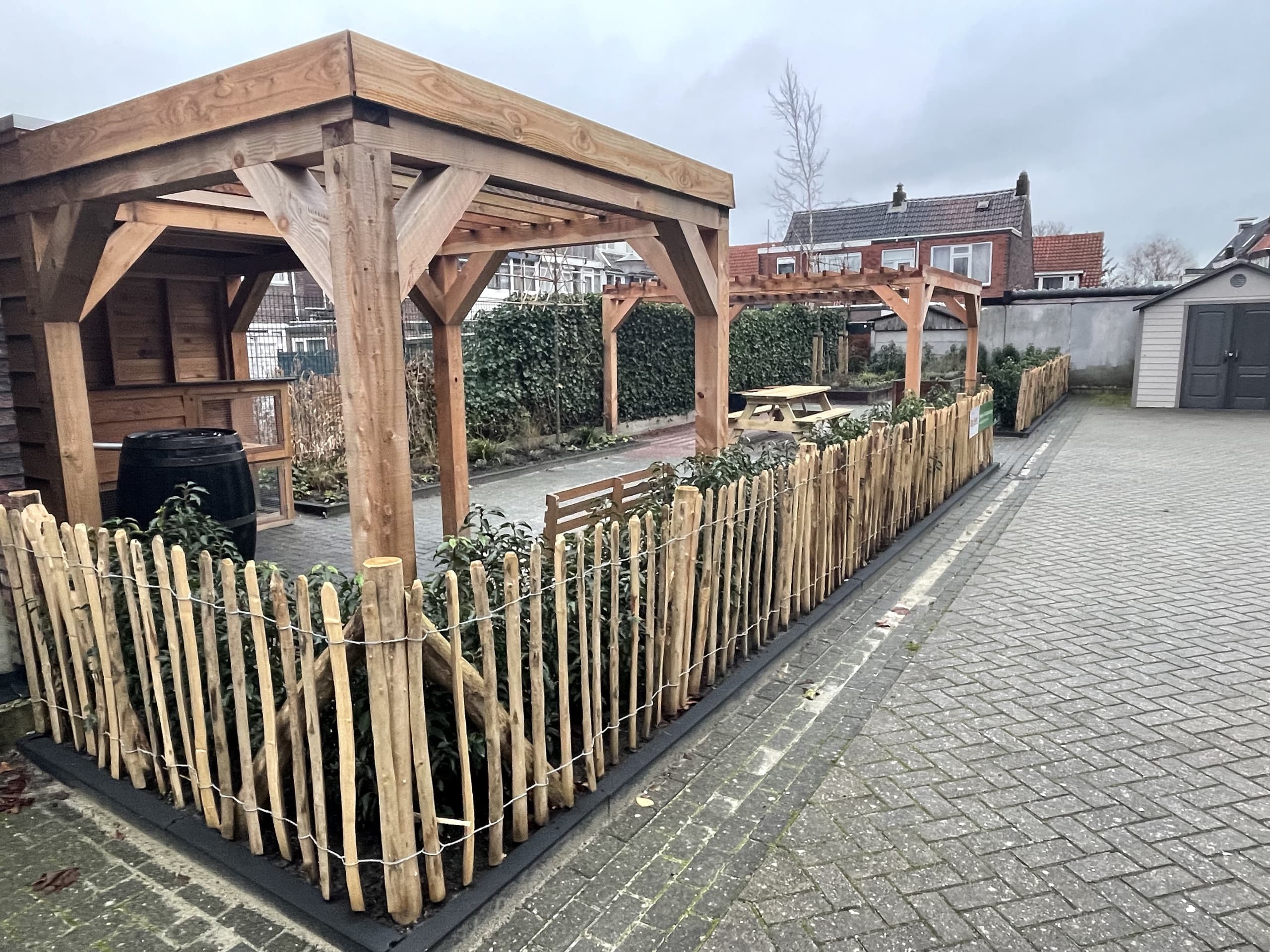De pergola en een overkapping gezien vanaf de bestrating, met de nieuwe hazelaar-erfafscheiding en jonge haag op de voorgrond