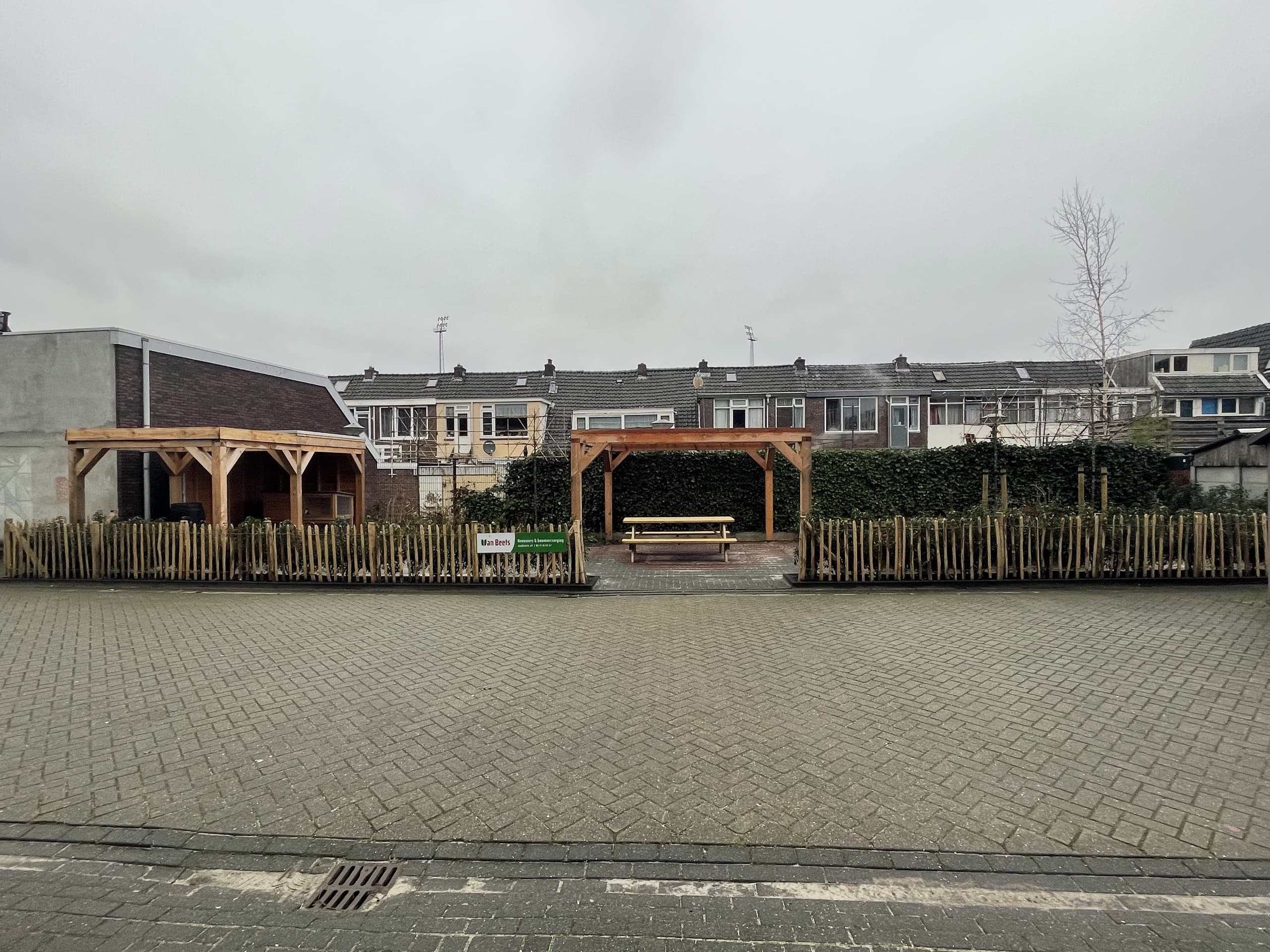 De tuin in de volle breedte, met het Van Beets-bord bij de picknicktafel onder de pergola en de rijtjeshuizen erachter