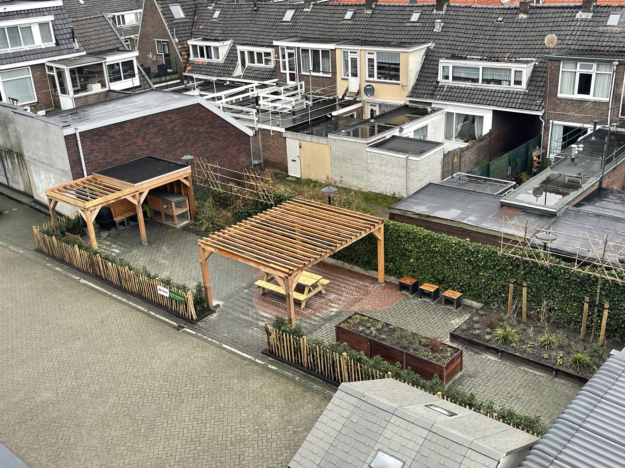 Luchtfoto van de volledige tuin: twee houten overkappingen, een pergola boven een picknicktafel, moestuinbakken en de hazelaar-erfafscheiding