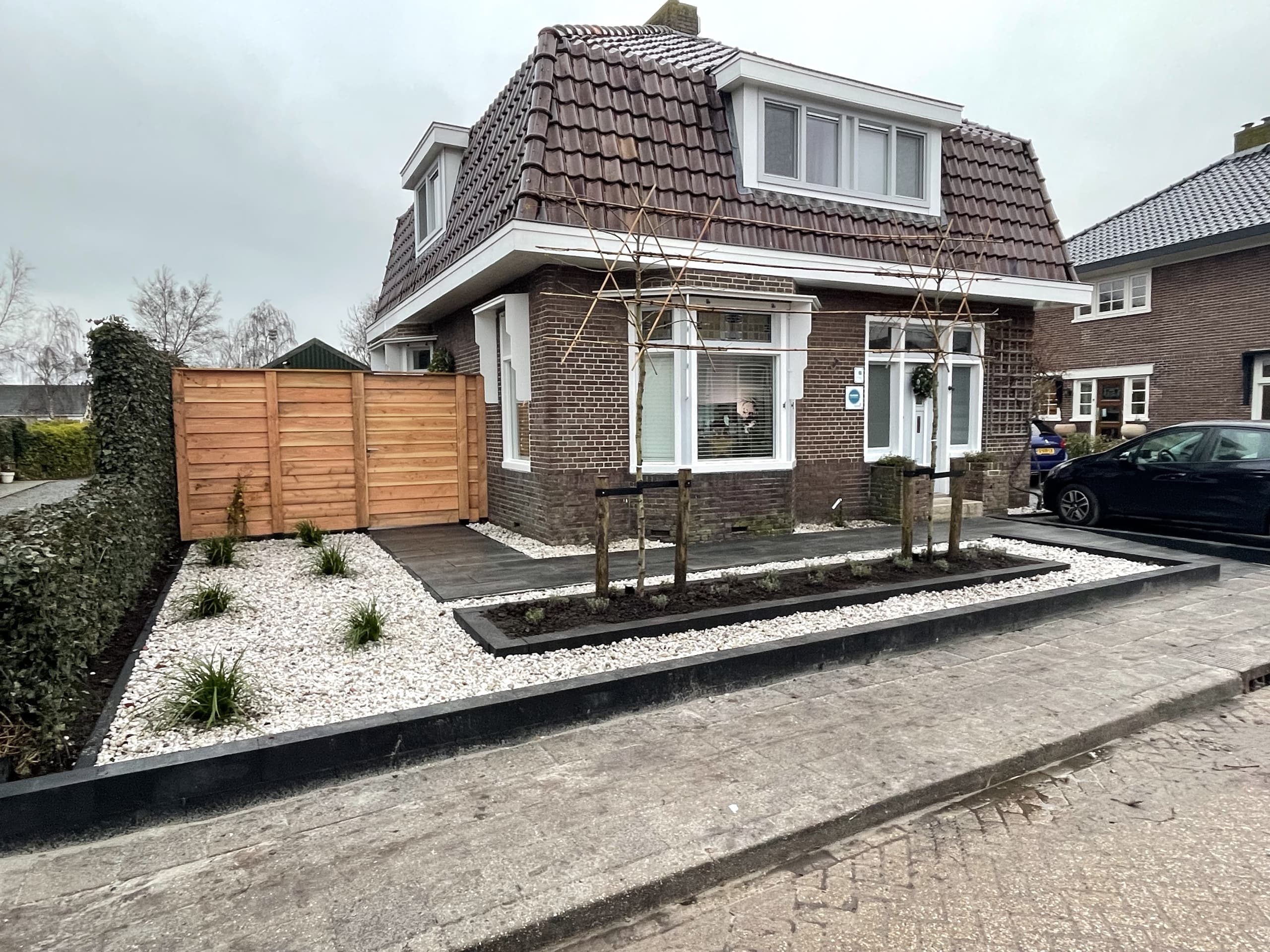 De nieuwe voortuin met wit split, siergrassen en twee jonge meerstammige bomen, met een houten poort naast de oprit
