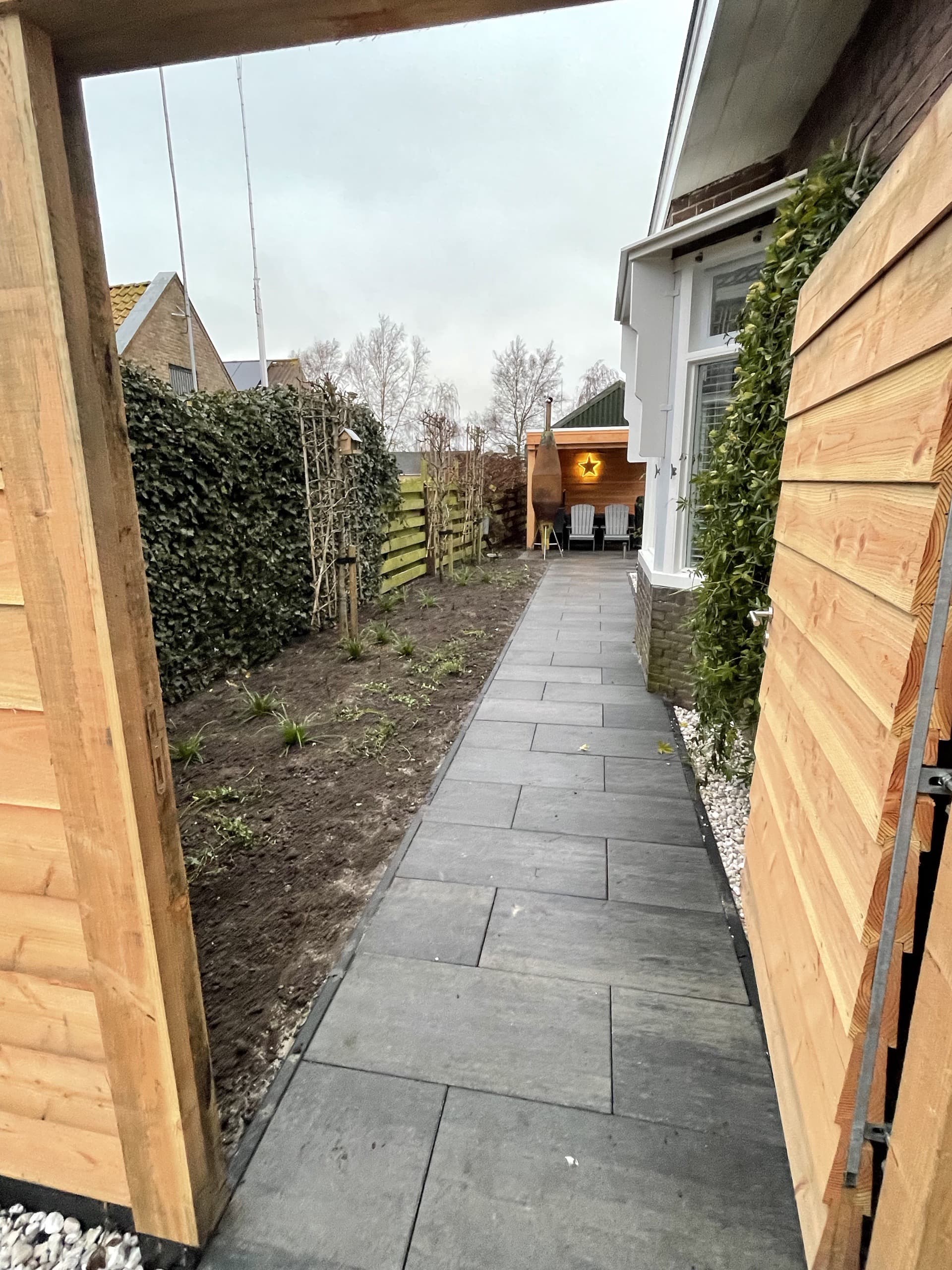 Het nieuwe tegelpad langs de border met leibomen op stok, vanaf de houten poort naar de overkapping achterin