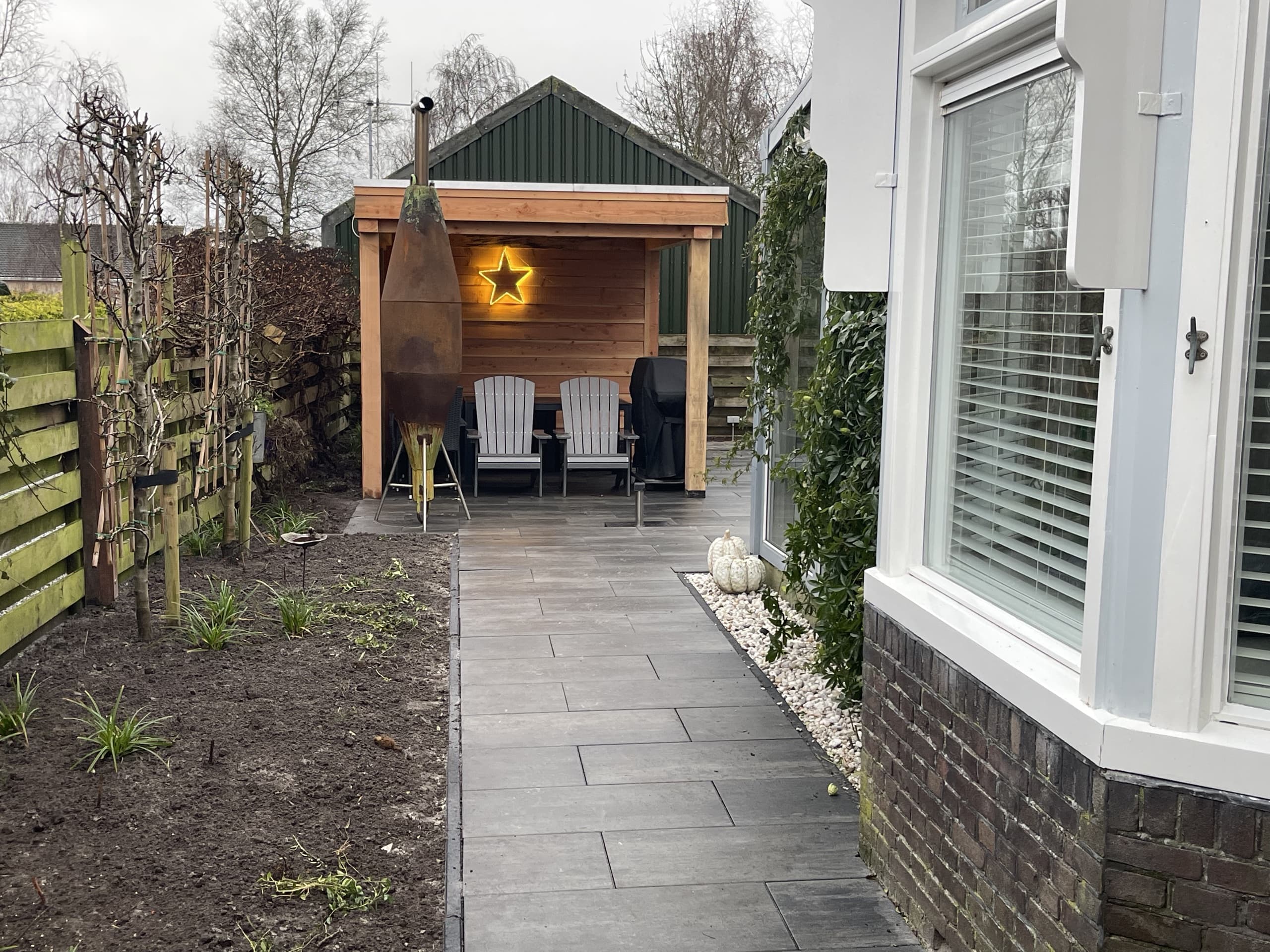 De houten overkapping met loungestoelen en een cortenstalen terrasheater, met de border ervoor nog in aanplant