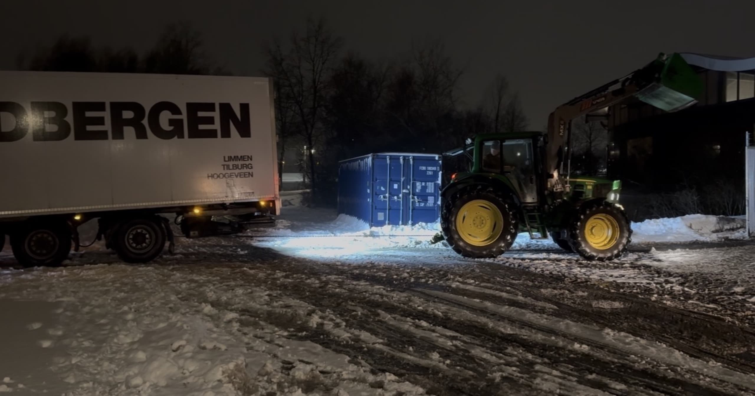 Tractor trekt in het donker met een sleepkabel een vastgereden vrachtwagentrailer los op een besneeuwd bedrijfsterrein