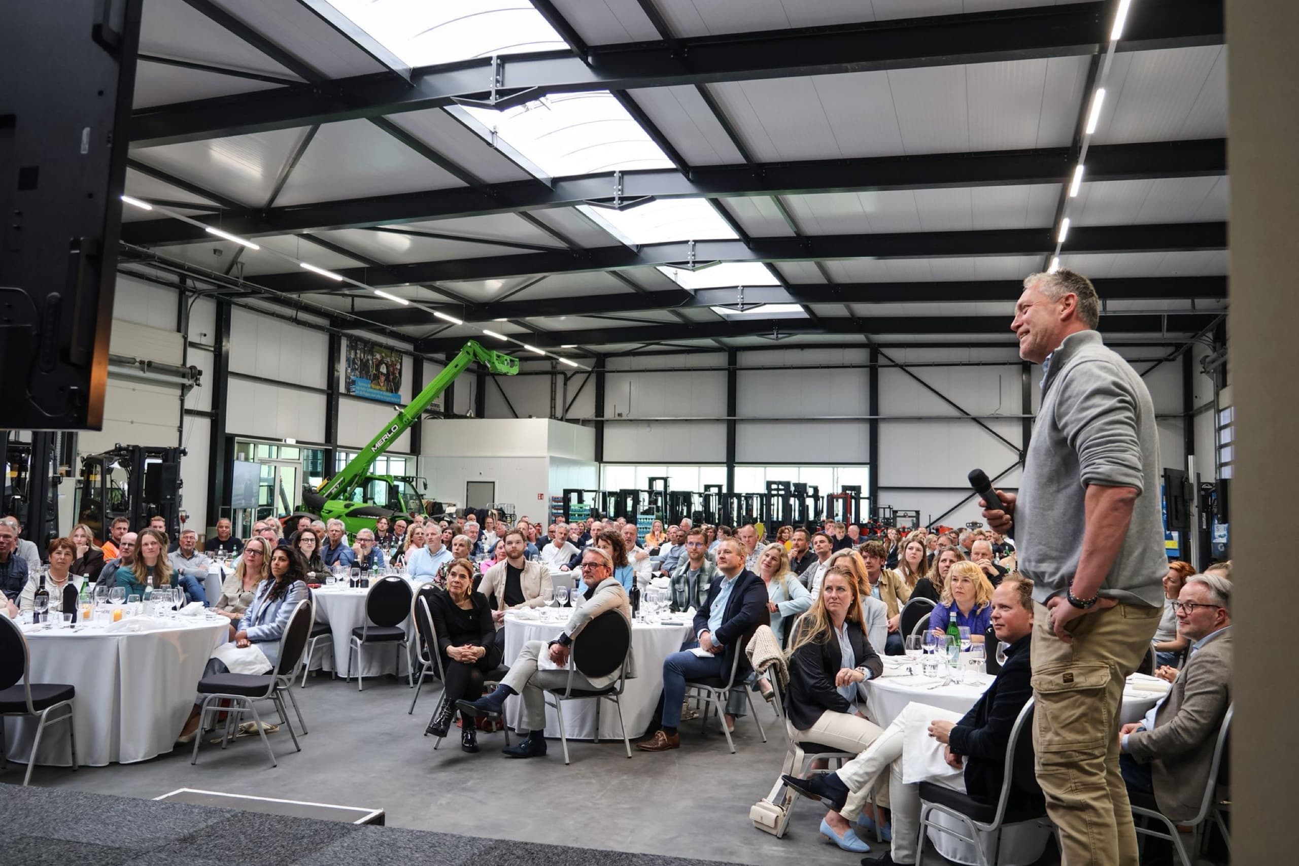Meine van Beets staat met een microfoon in de hand op het podium en spreekt een volle zaal toe in een bedrijfshal met gedekte tafels