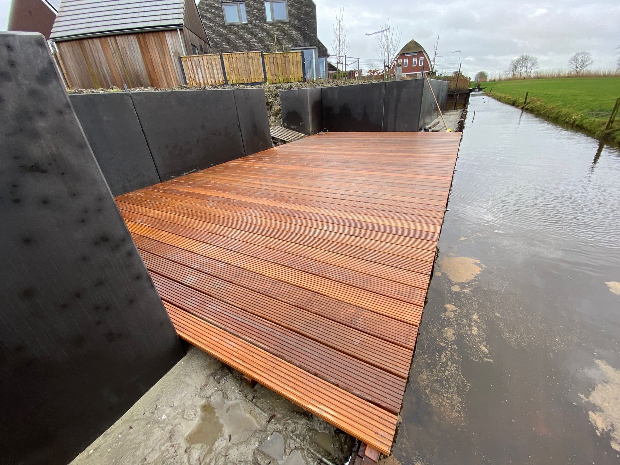 Nieuwe steiger van hardhout in een sloot, tussen zwarte keerwanden bij nieuwbouwwoningen