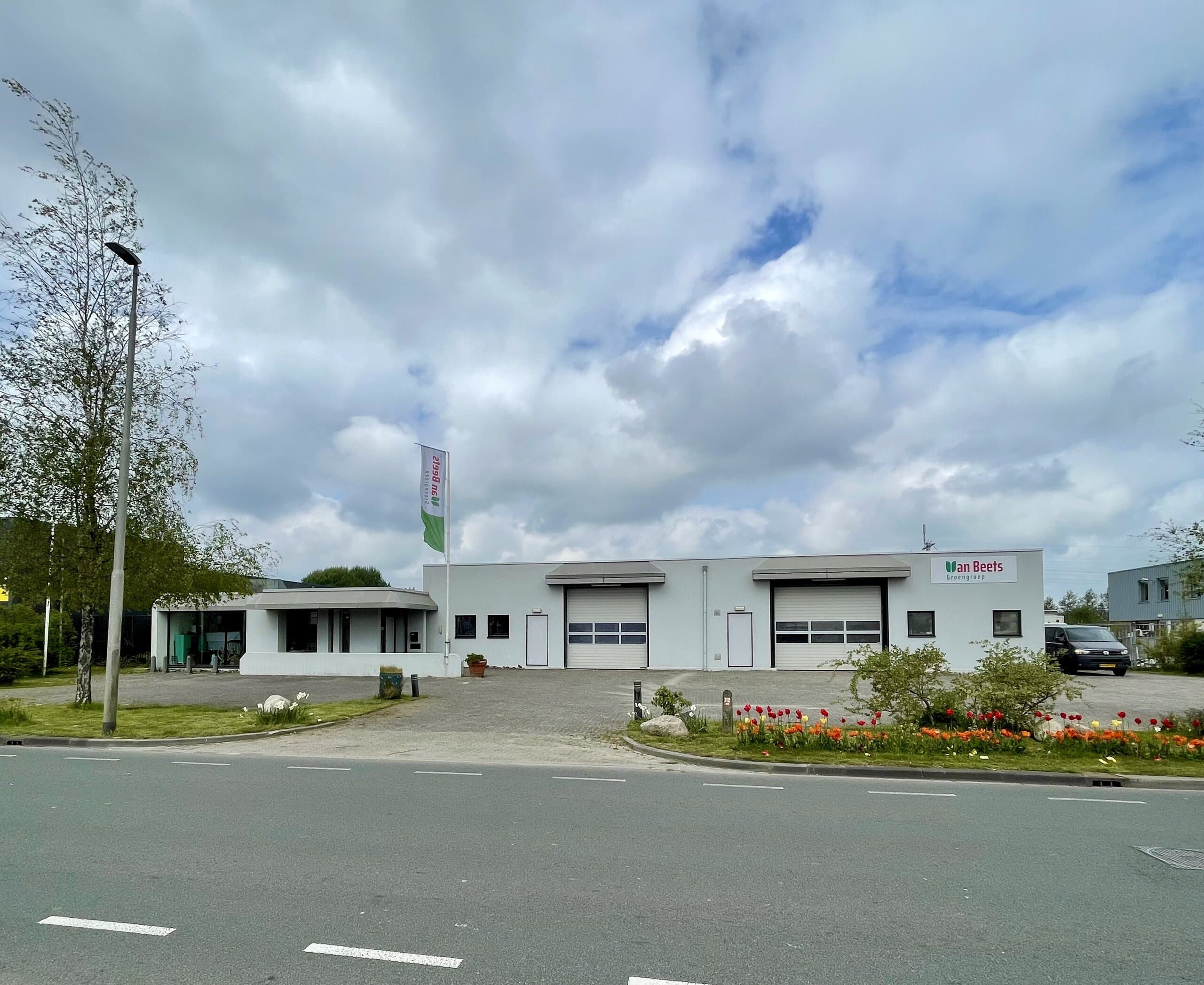 Het bedrijfspand van Van Beets Groengroep aan de Uranusweg in Leeuwarden: een licht bedrijfsgebouw met twee roldeuren, het logo op de gevel, een vlag met de bedrijfsnaam en een border met bloeiende tulpen langs de stoep