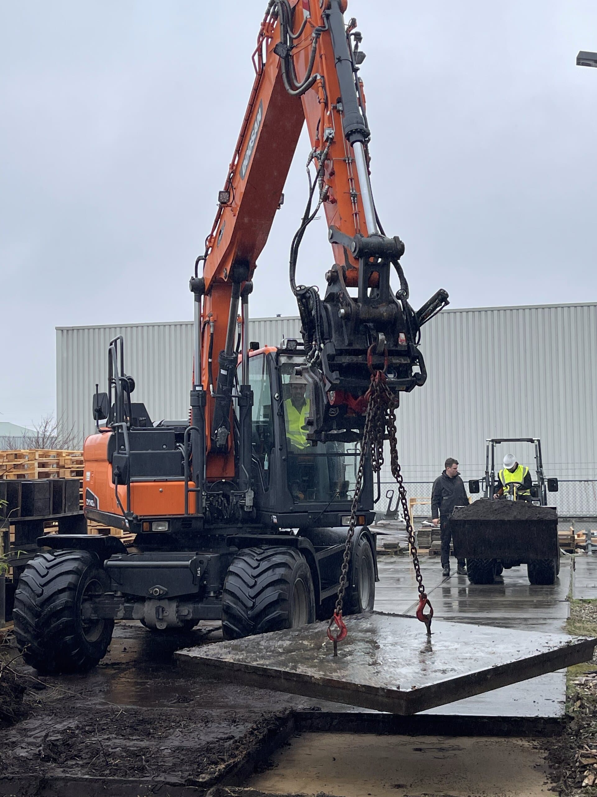 Een graafmachine tilt met kettingen een betonplaat uit het terrein tijdens een natte werkdag