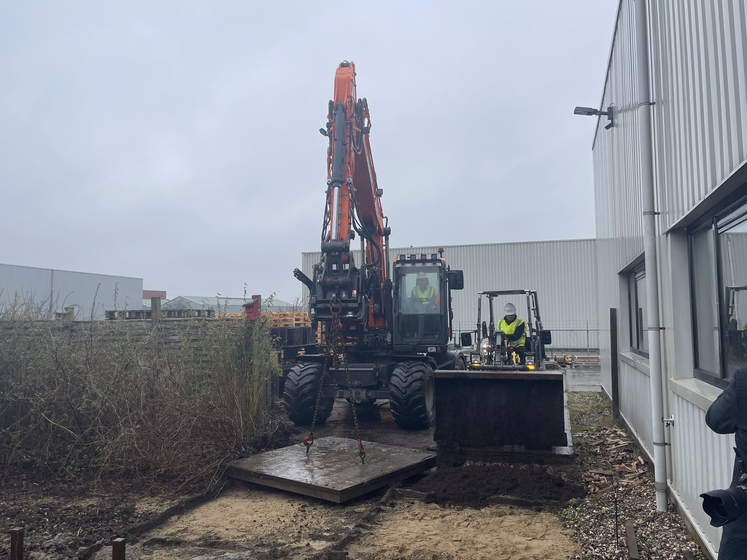 Rupskraan tilt een betonplaat weg terwijl een collega met de shovel de vrijgekomen grond naast het bedrijfspand afvoert