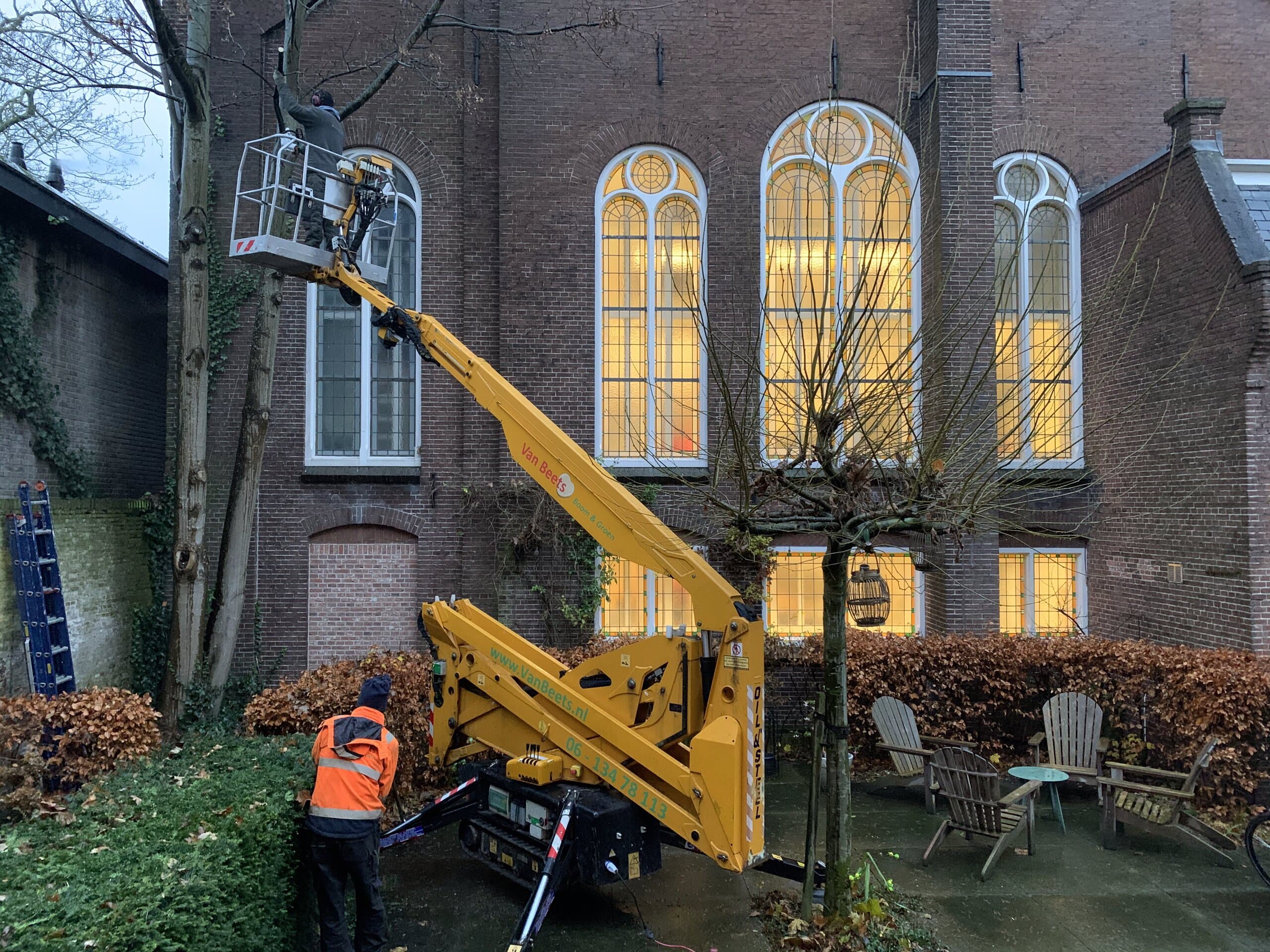 Boomverzorger van Van Beets snoeit vanuit een rupshoogwerker een boom naast een kerkgebouw, terwijl een collega het werk vanaf de grond begeleidt