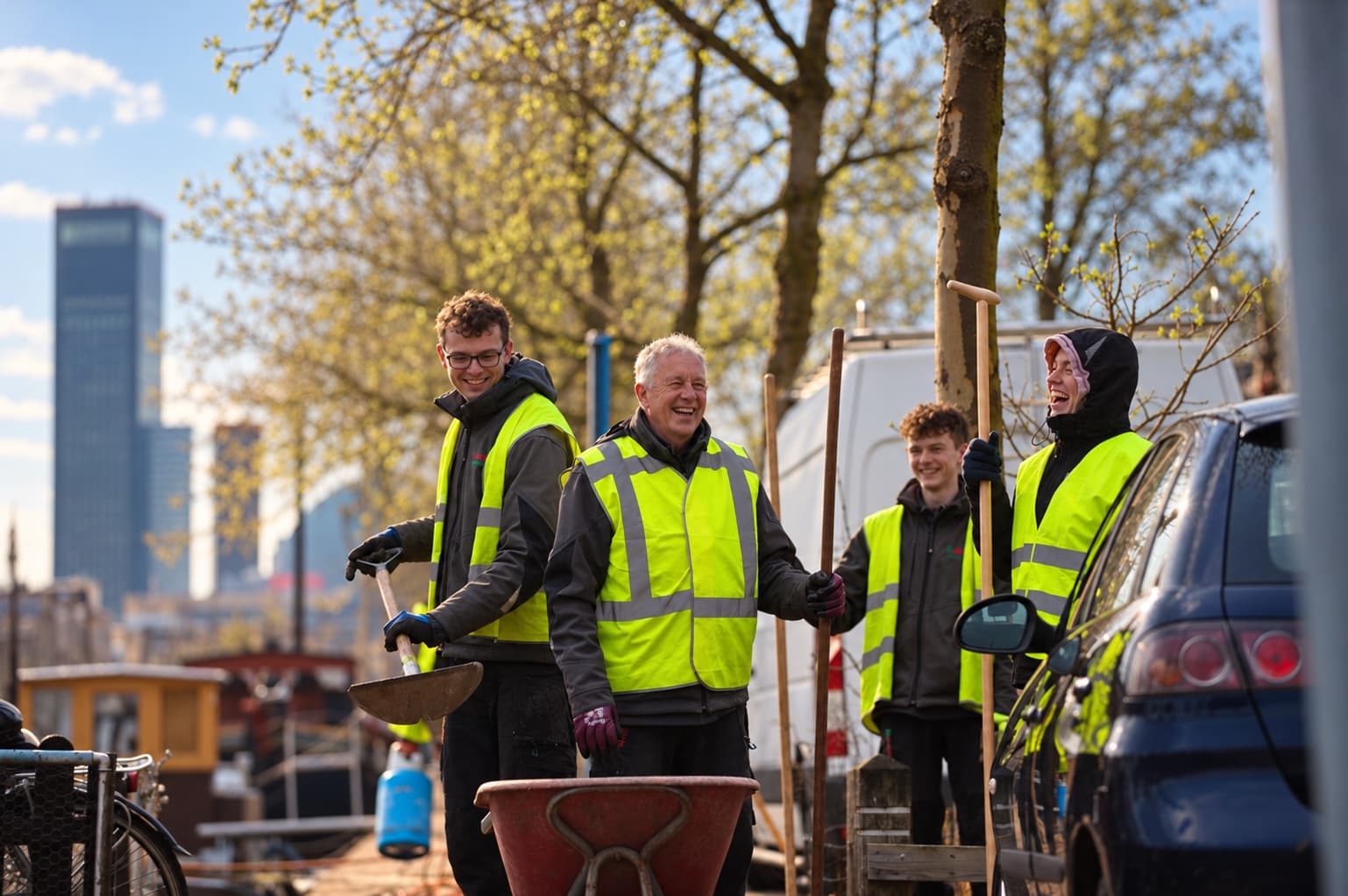 Vier collega's van Van Beets, lachend aan het werk op de kades in Leeuwarden, in werkkleding met geel hesje, met een schep, boomstaken en een kruiwagen bij de hand; op de achtergrond woonboten en de hoogbouw van de stad. Deze foto is met AI bewerkt.