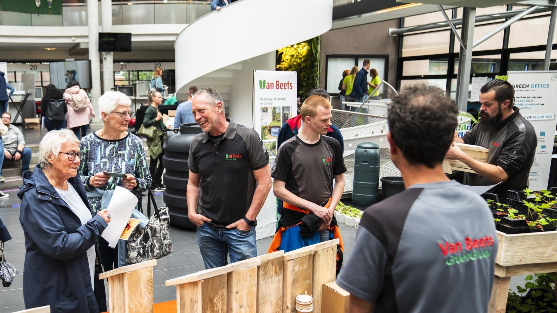 Collega's van Van Beets in bedrijfskleding staan in de hal van een gemeentehuis bij een tafel met zelfgemaakte vogelhuisjes en praten met bezoekers
