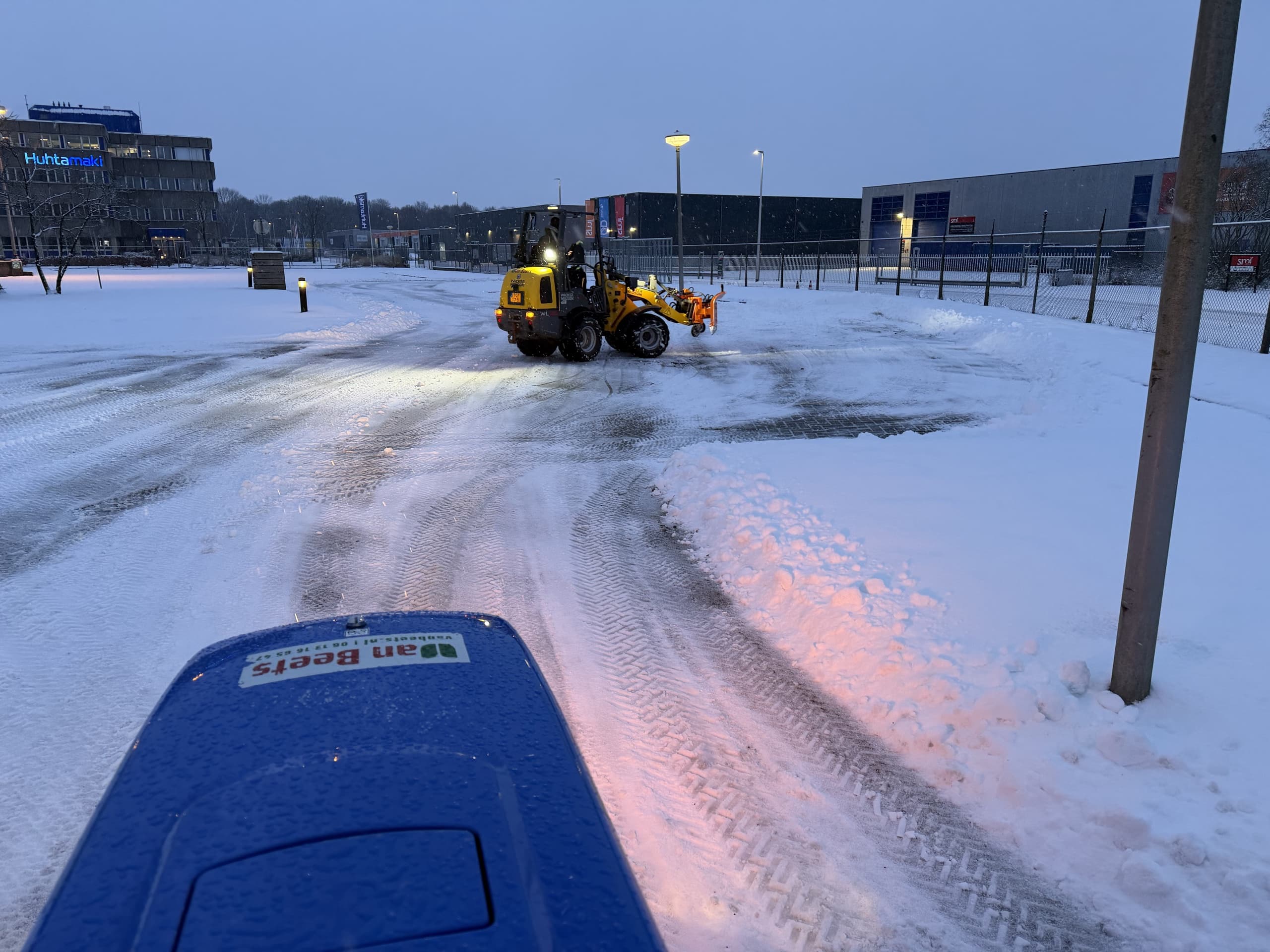 Wiellader met sneeuwschuif ruimt bij schemering een besneeuwd bedrijventerrein tussen kantoorpanden, met de blauwe bus van Van Beets op de voorgrond