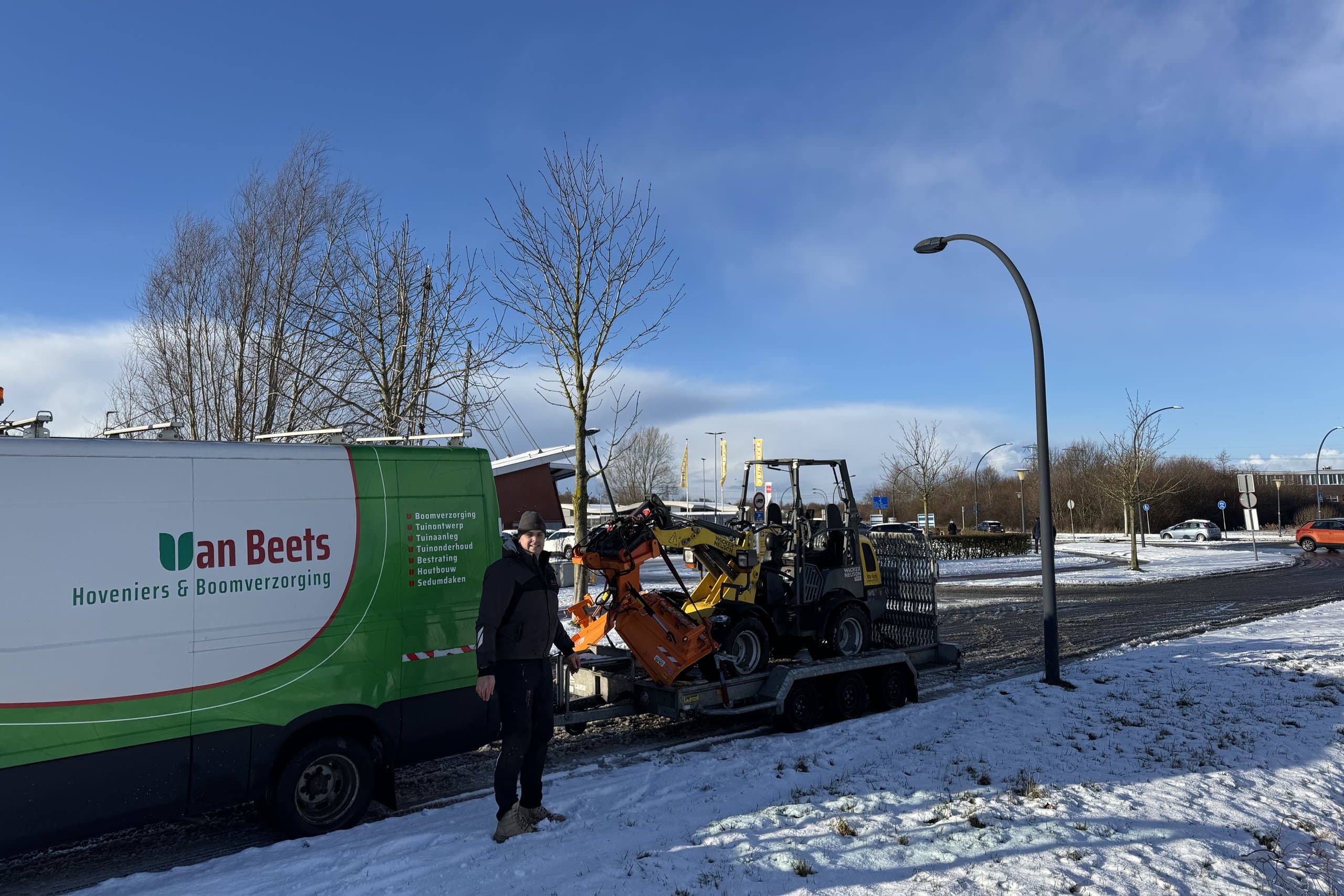 Een medewerker van Van Beets bij de bus en een aanhanger met een kleine shovel op een besneeuwd bedrijventerrein