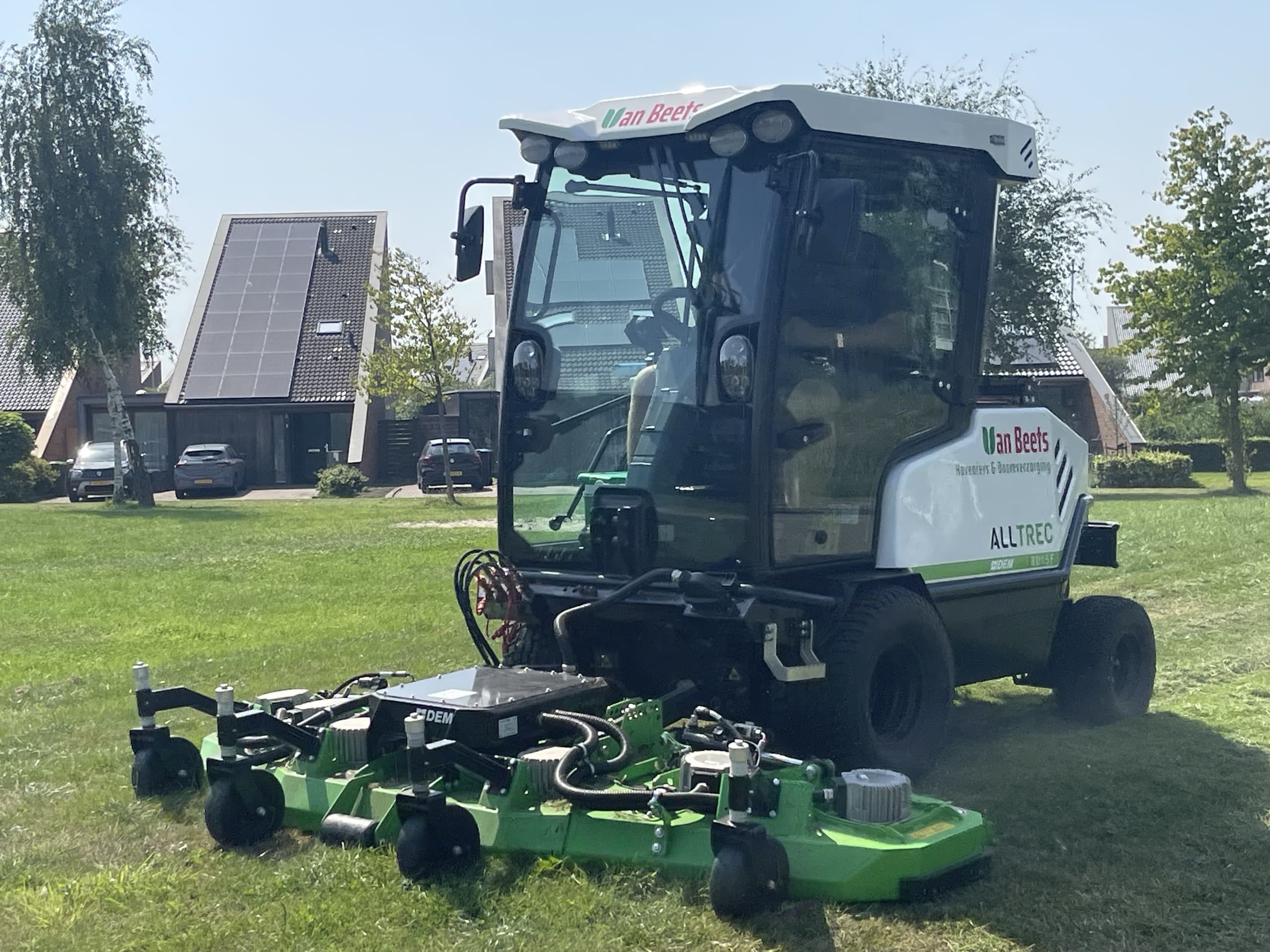 De elektrische maaimachine van Van Beets van dichtbij, met de cabine en het brede maaidek, op een gazon tussen de woningen