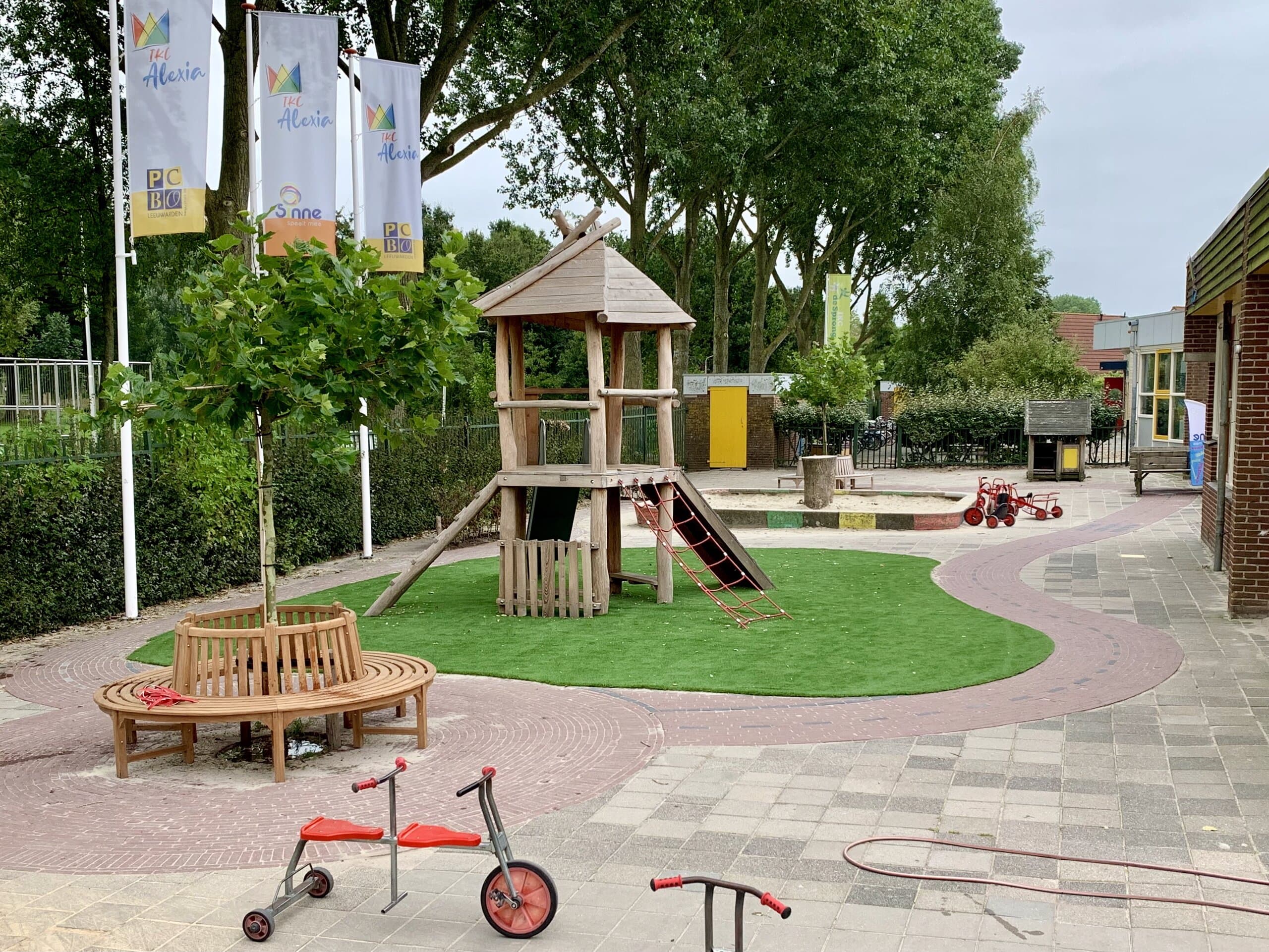 Aangelegd schoolplein met speeltoestel, kunstgras, boombank en sierbestrating in een gebogen patroon