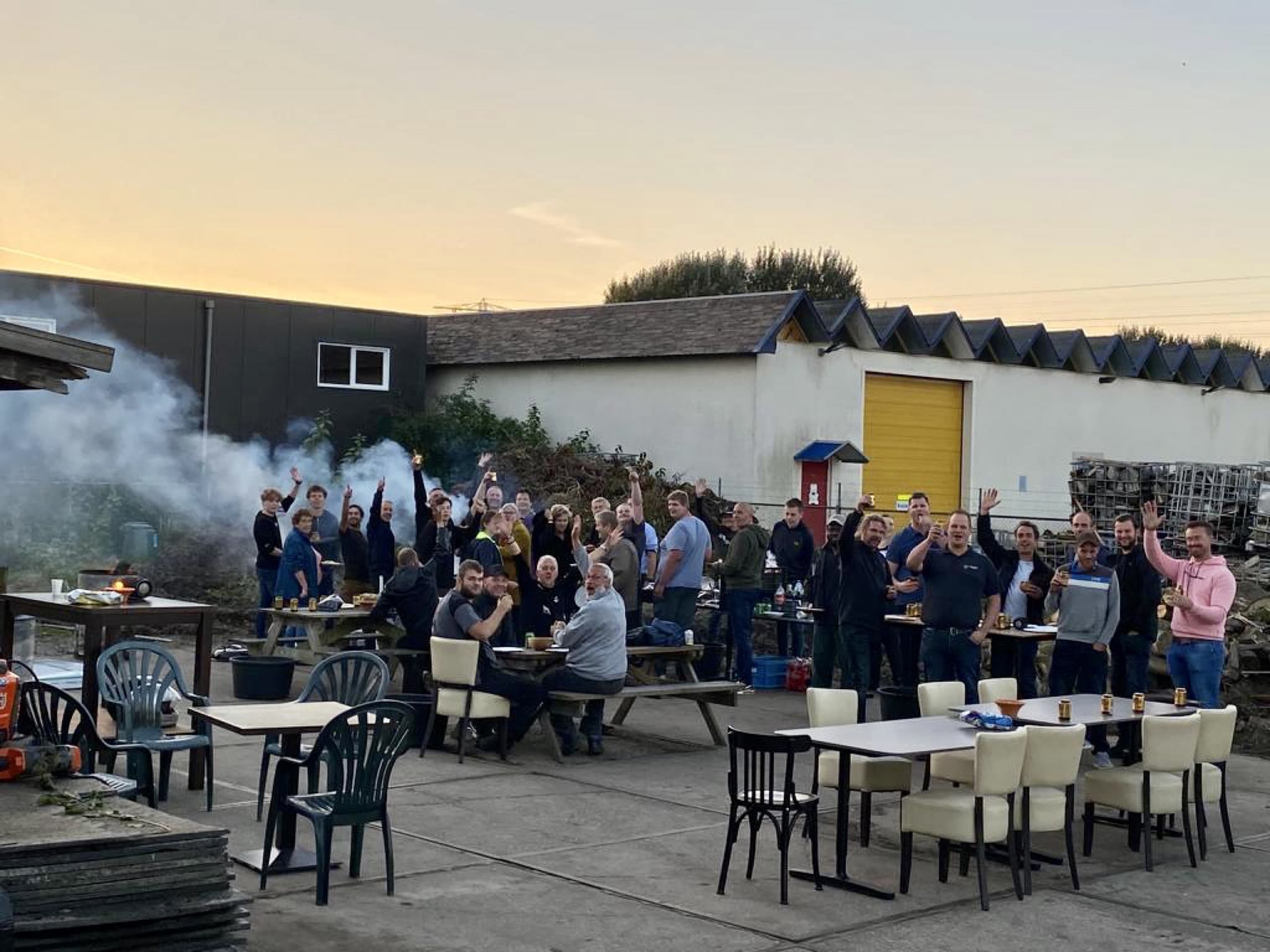 Het team van Van Beets zwaait naar de camera op het buitenterrein bij de loods, rond een paar tafels met de barbecue op de achtergrond