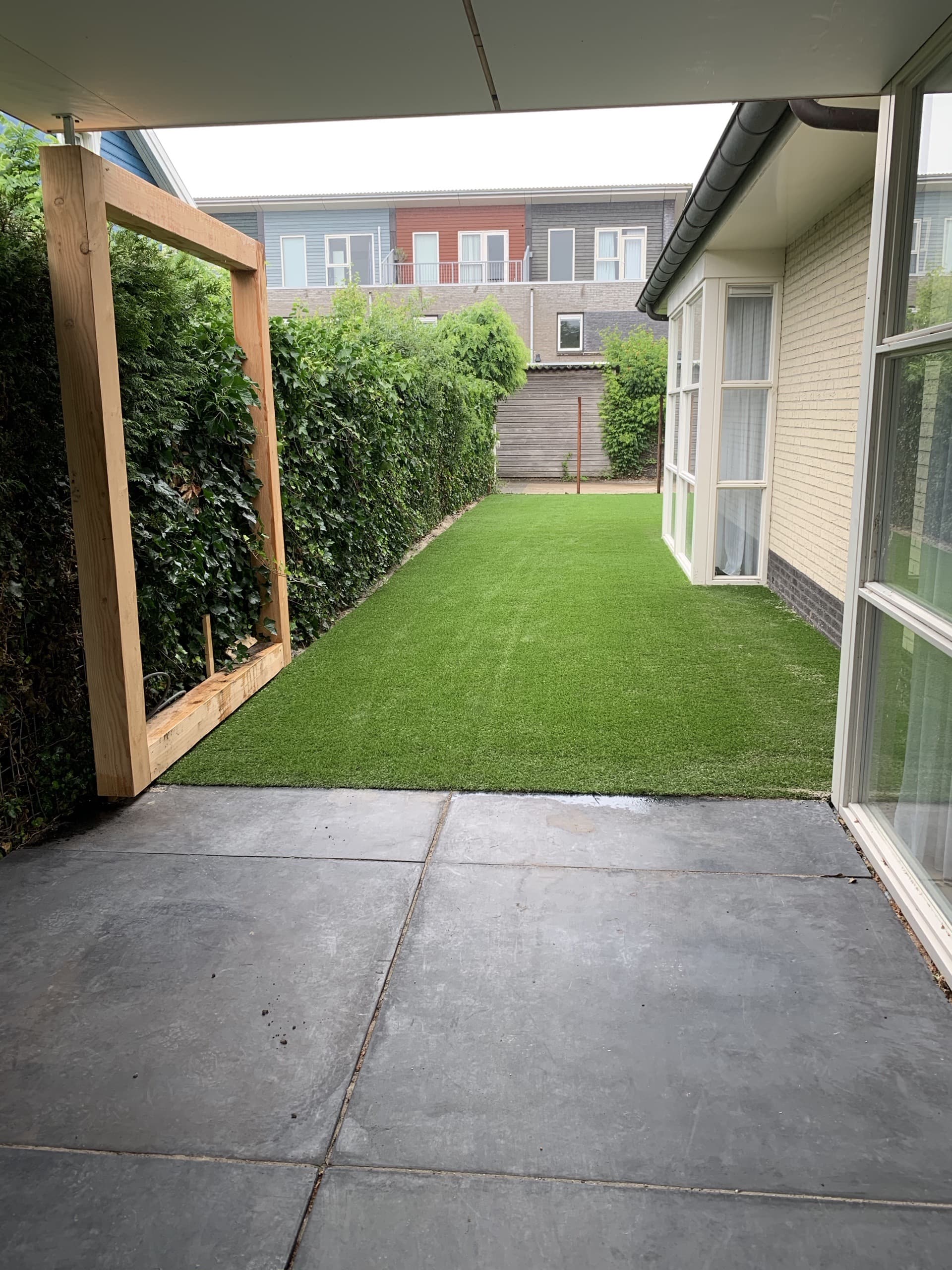 Kunstgras tussen het huis en de klimopwand, met een houten frame dat de begroeiing draagt