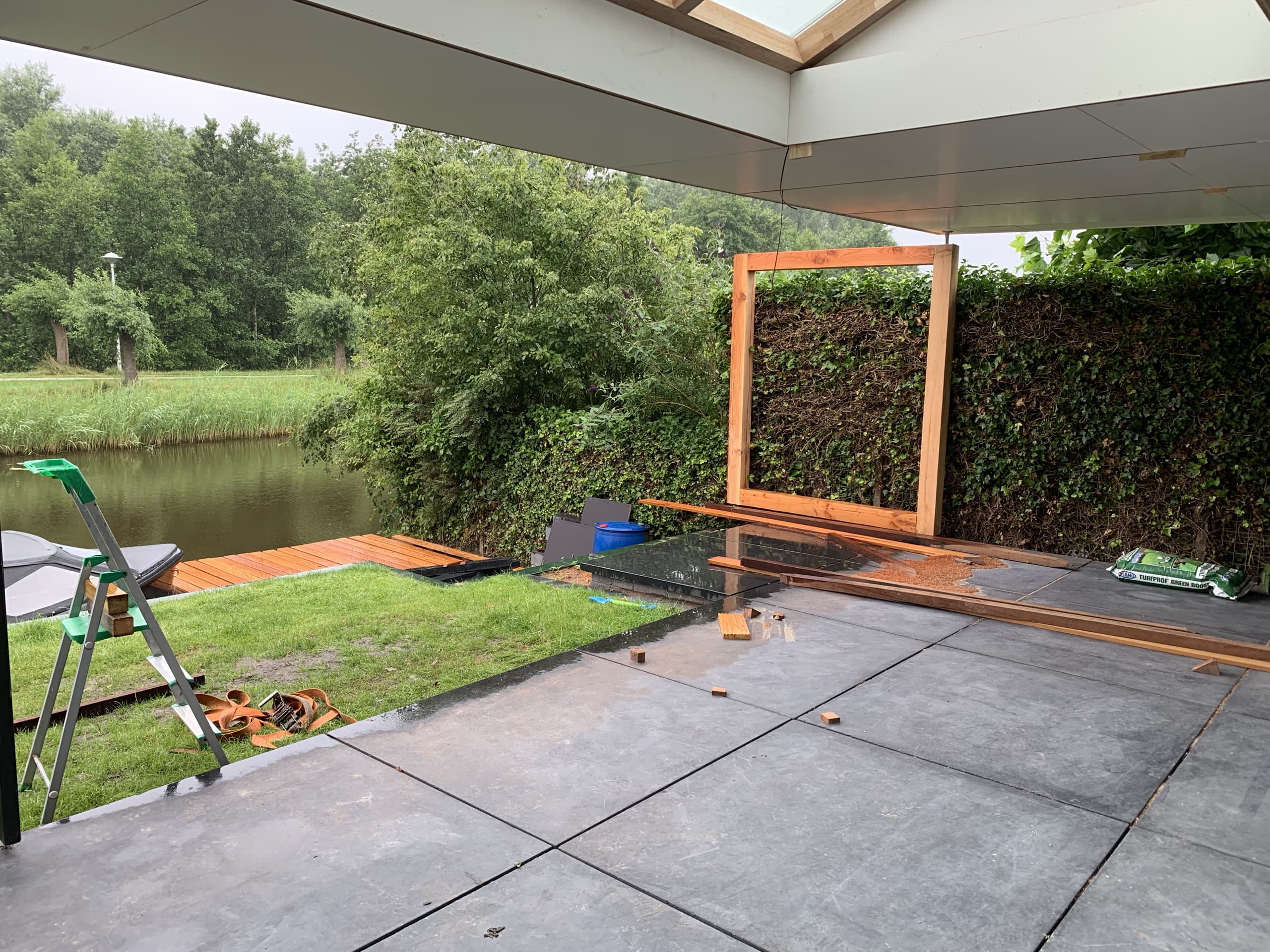 Het terras in aanleg: grote grijze tegels lopen door tot aan de vlonder aan het water, met een houten frame tegen de klimopwand