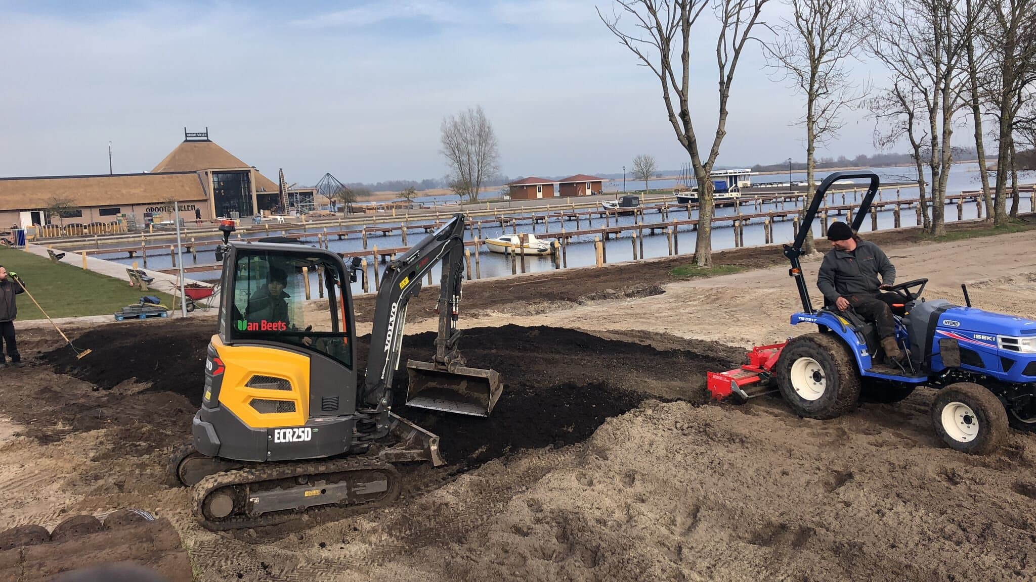 Minigraver en tractor met grondfrees maken de ondergrond klaar langs een aanlegsteiger, terwijl een collega de zwarte grond aanharkt