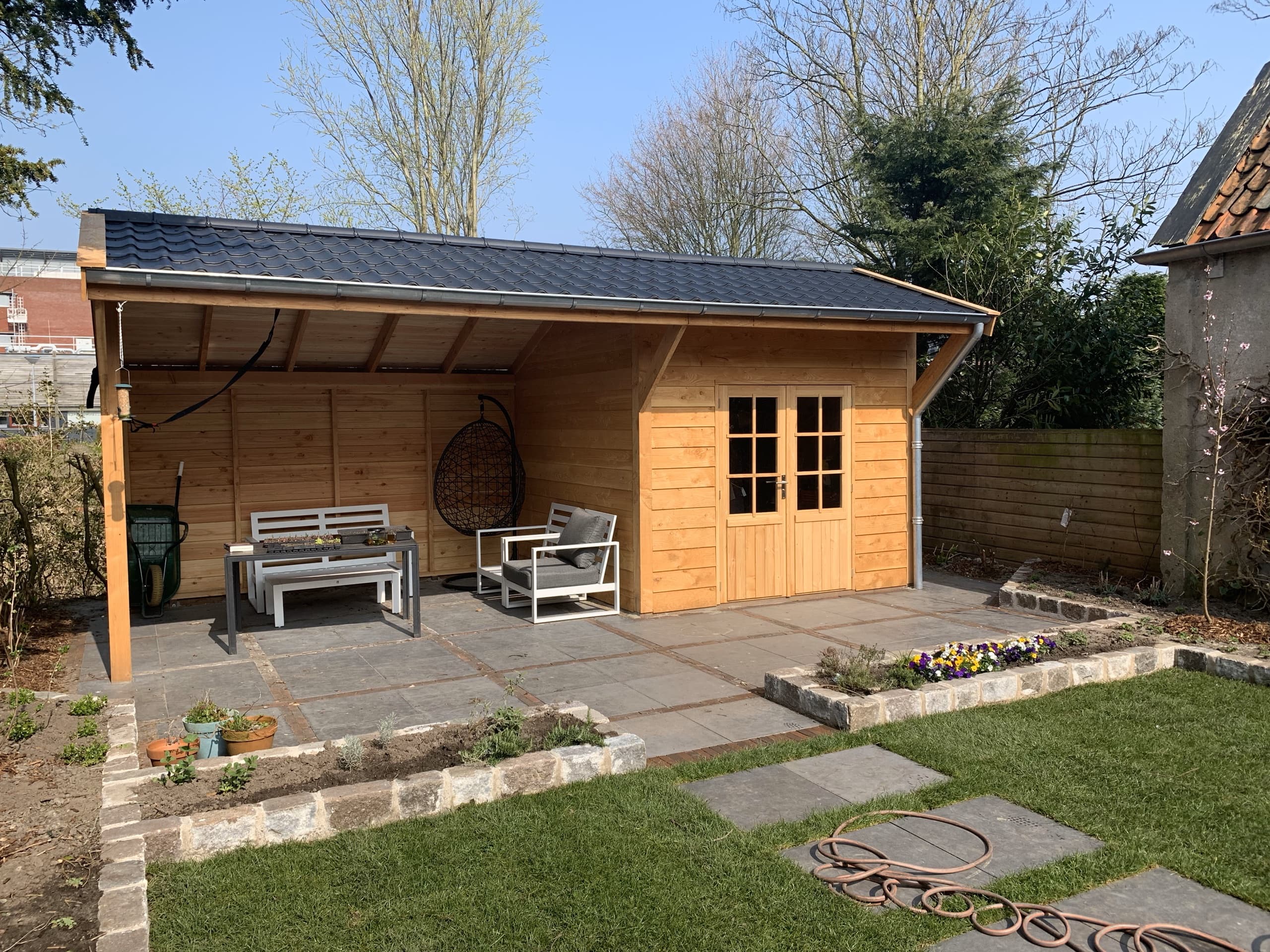 Houten tuinhuis met een overkapping ervoor, ingericht als zithoek met loungestoelen en een hangstoel
