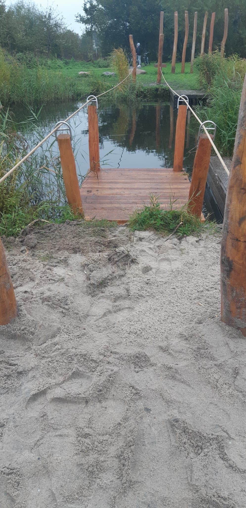 Houten aanlegsteiger met bolpalen en touwleuning aan het water, met een zandpad ervoor